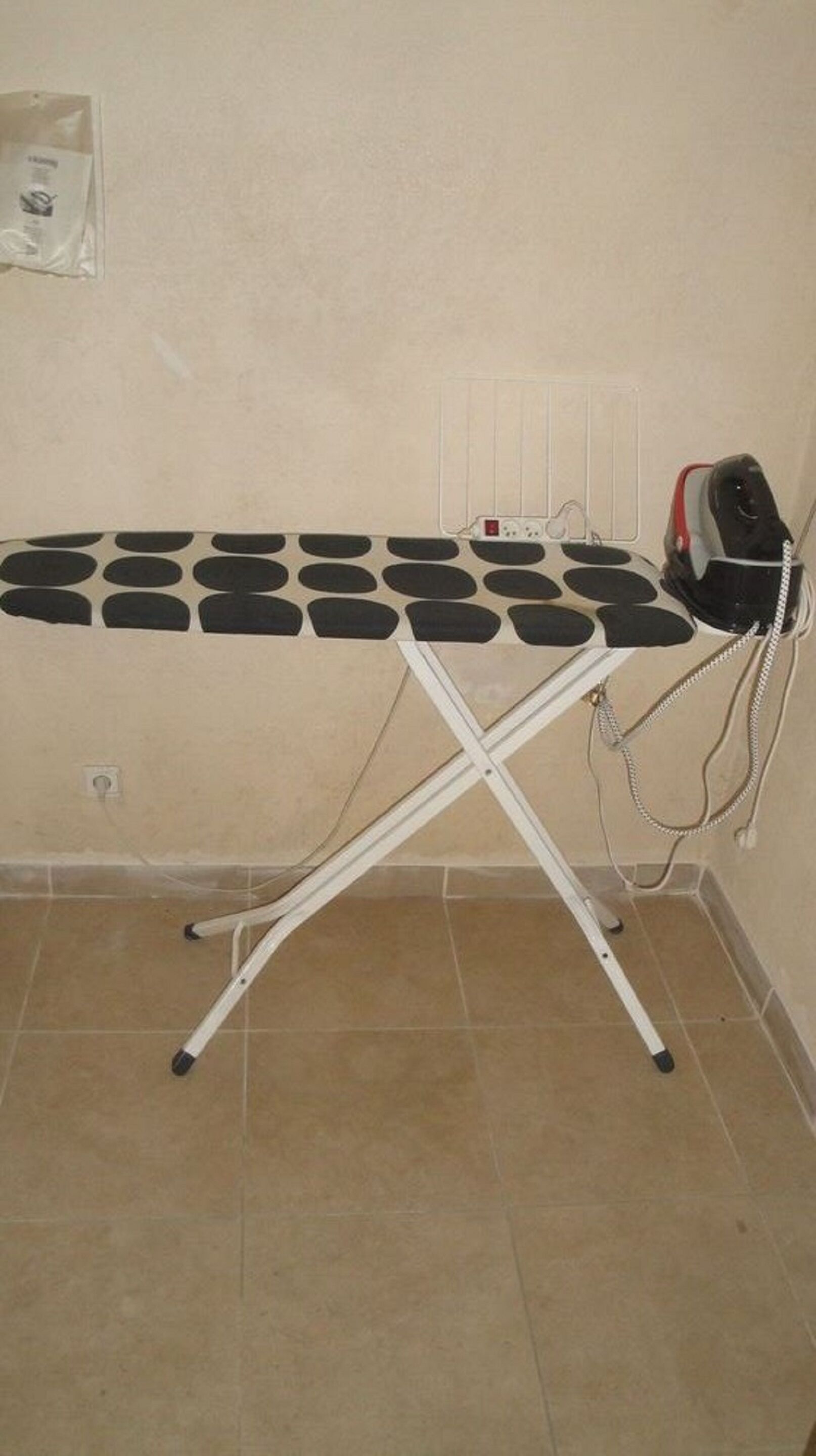 Una tabla de planchar con funda a lunares blanco y negro, un hierro rojo y negro, y un enchufe en la pared.