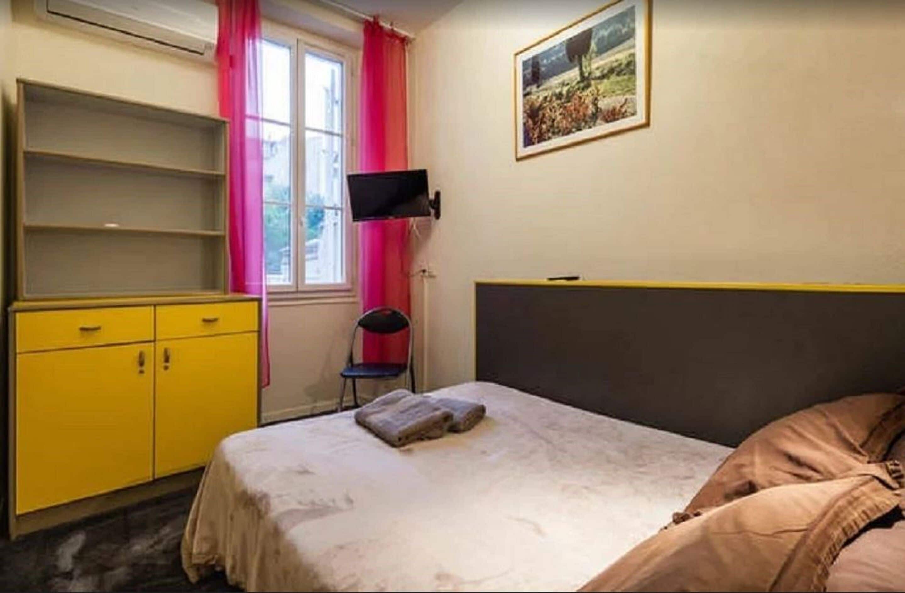Une chambre à coucher avec un lit, une armoire jaune, un téléviseur fixé au mur et une fenêtre avec des rideaux roses.
