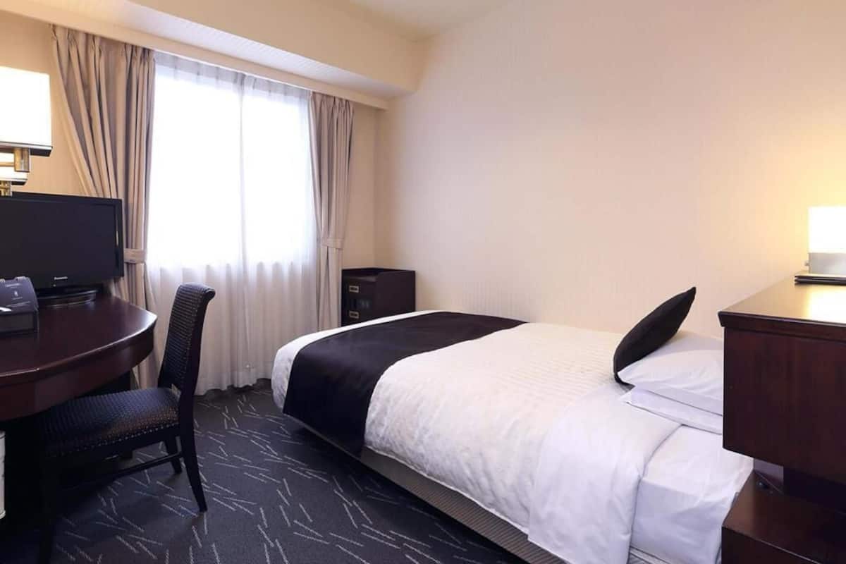 A hotel room with a bed, a desk with a chair, a television, and a window with curtains.