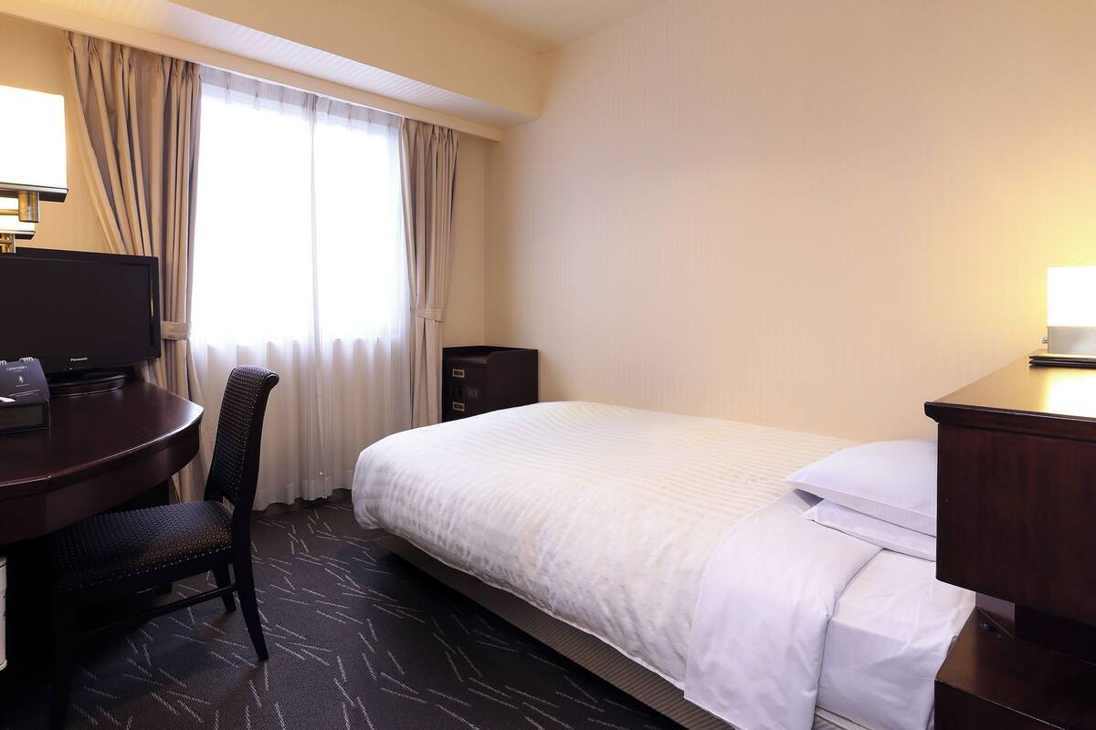 A hotel room with a bed, a desk with a television, a chair, and a nightstand.