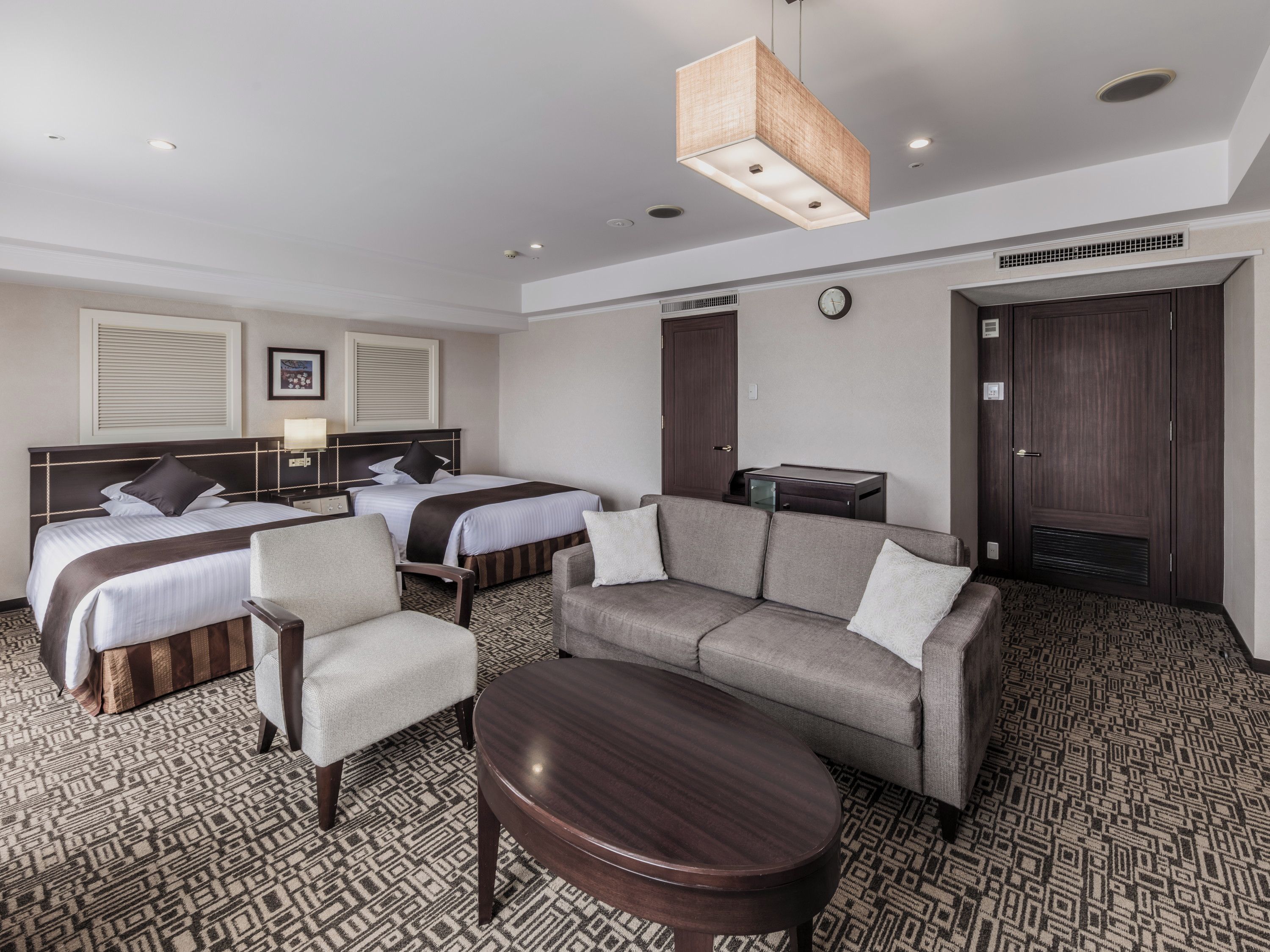 A hotel room with two beds, a sofa, a chair, a small table, and a clock on the wall.