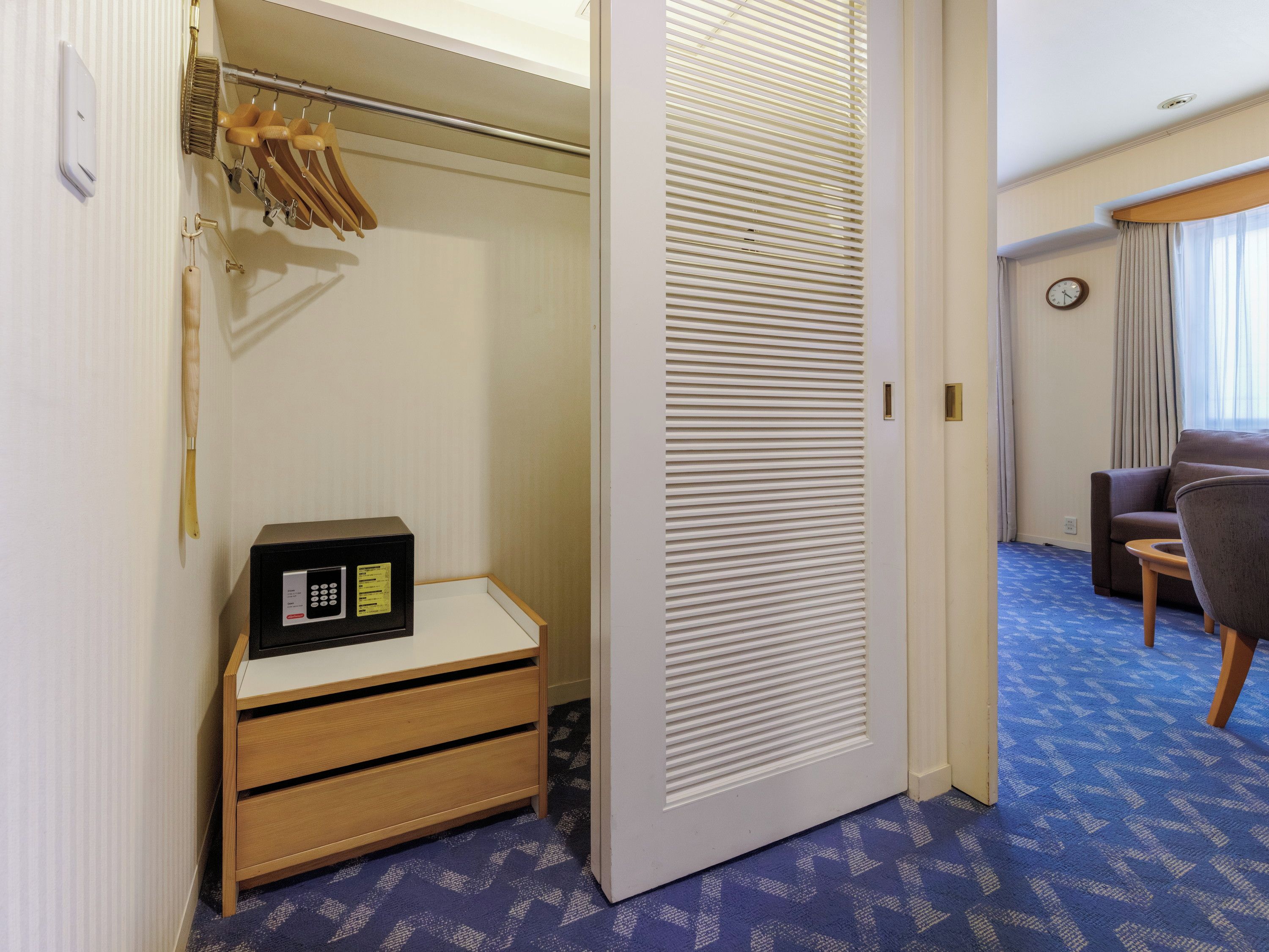 Ein Hotelzimmer mit begehbarem Kleiderschrank, einem kleinen Holzschrank, einem Bett mit Kopfteil, einer Sitzecke mit Sofa und Sesseln sowie einer Wanduhr.