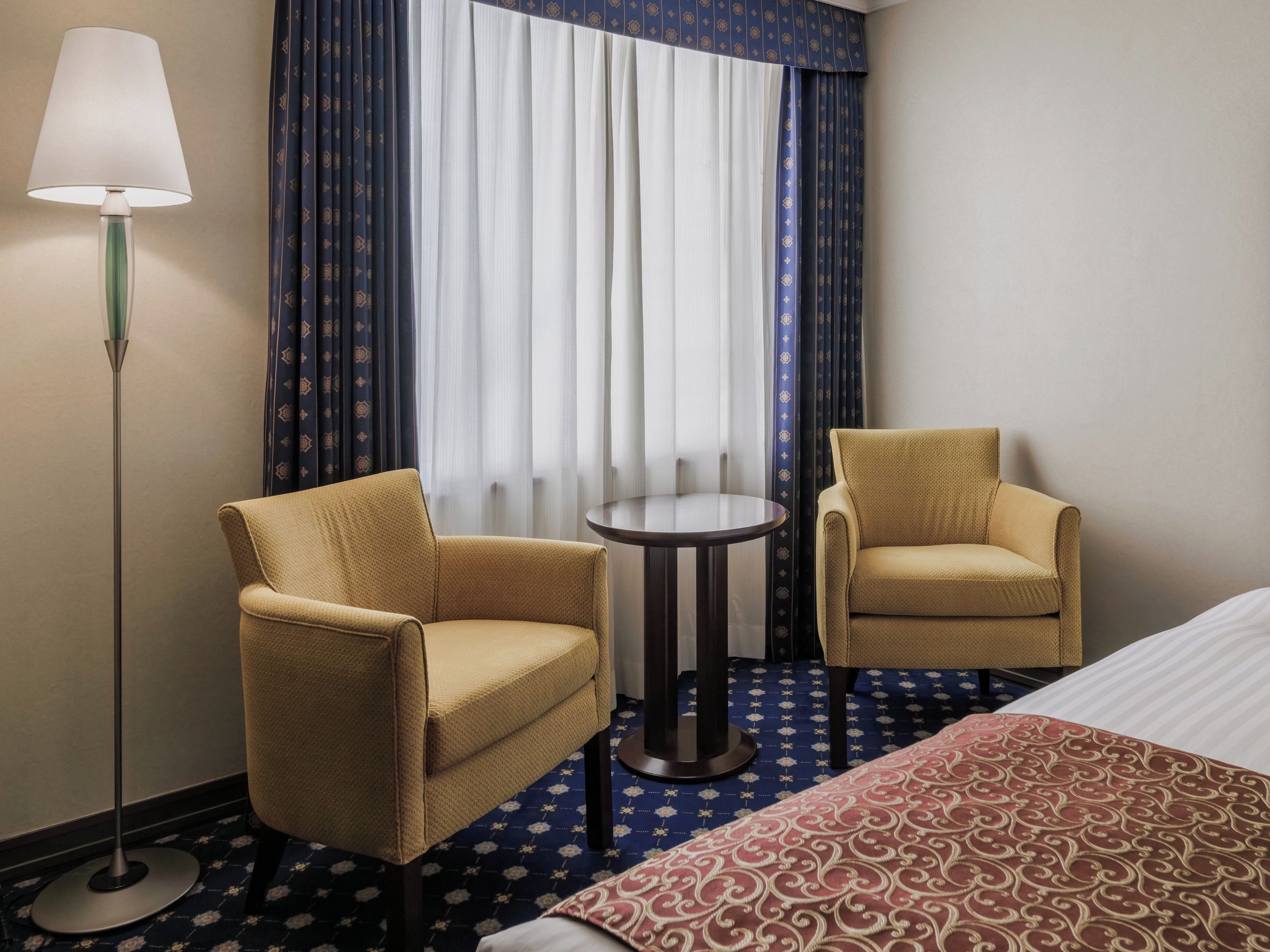 Ein Hotelzimmer mit zwei Sesseln, einem kleinen runden Tisch, einer Stehlampe und einem Bett mit einem gemusterten Überwurf.