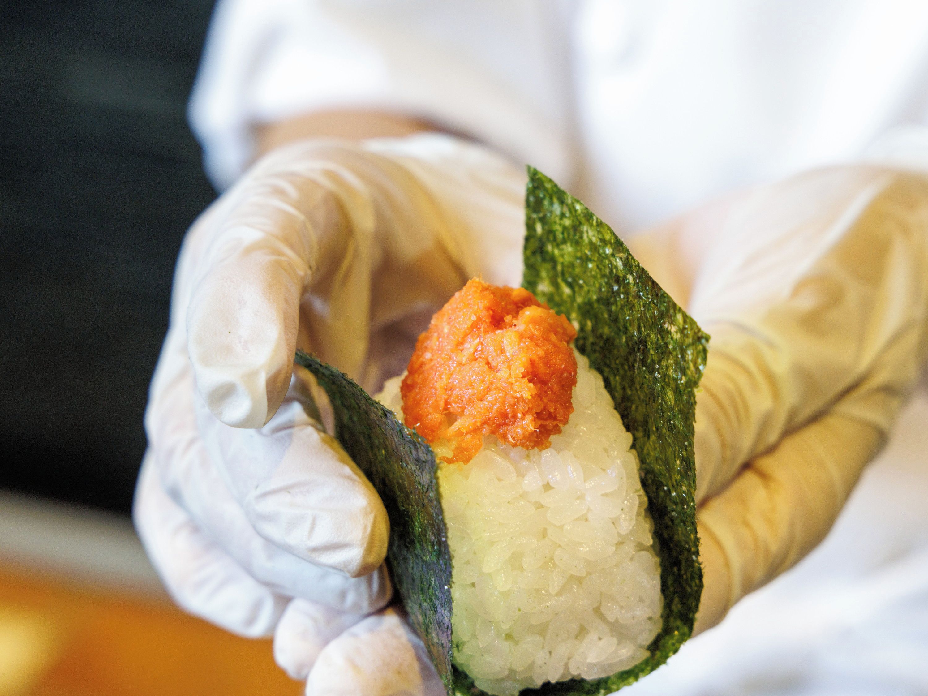 Mão com luva segurando um rolo de sushi com arroz e cobertura.