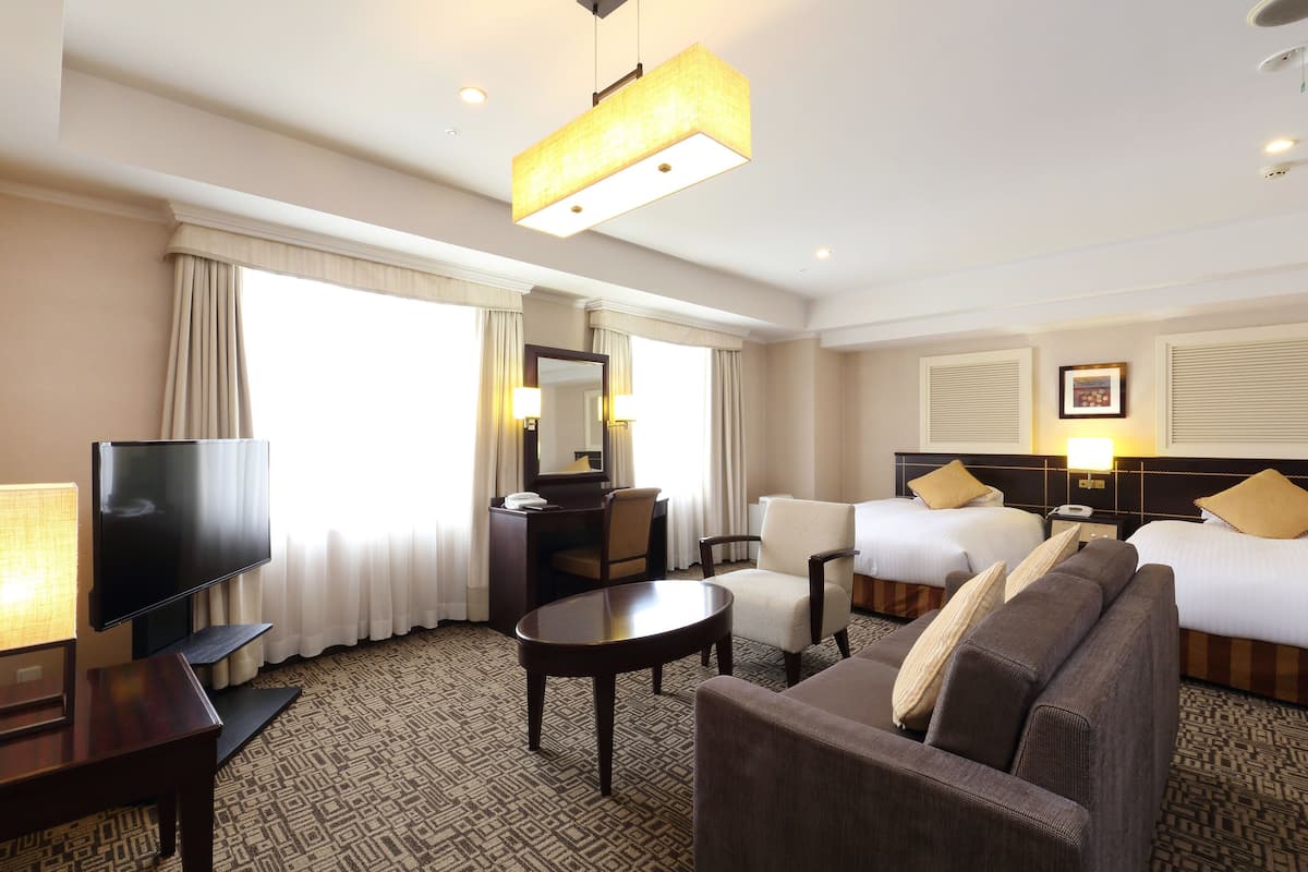 A hotel room with a television, two beds, a sofa, a desk, and a large window with curtains.