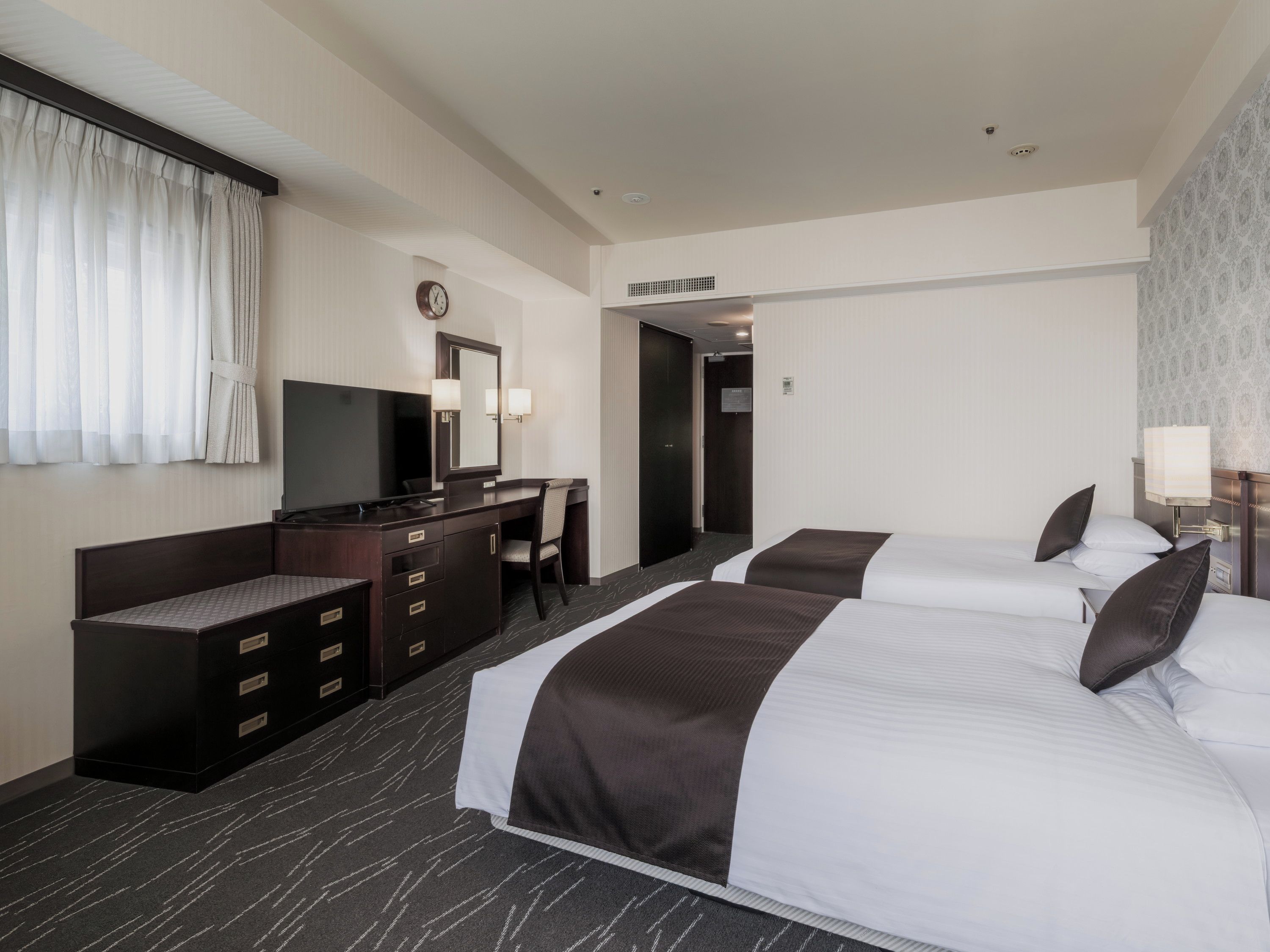 A hotel room with a bed, a desk, a chair, a television, and a clock on the wall.
