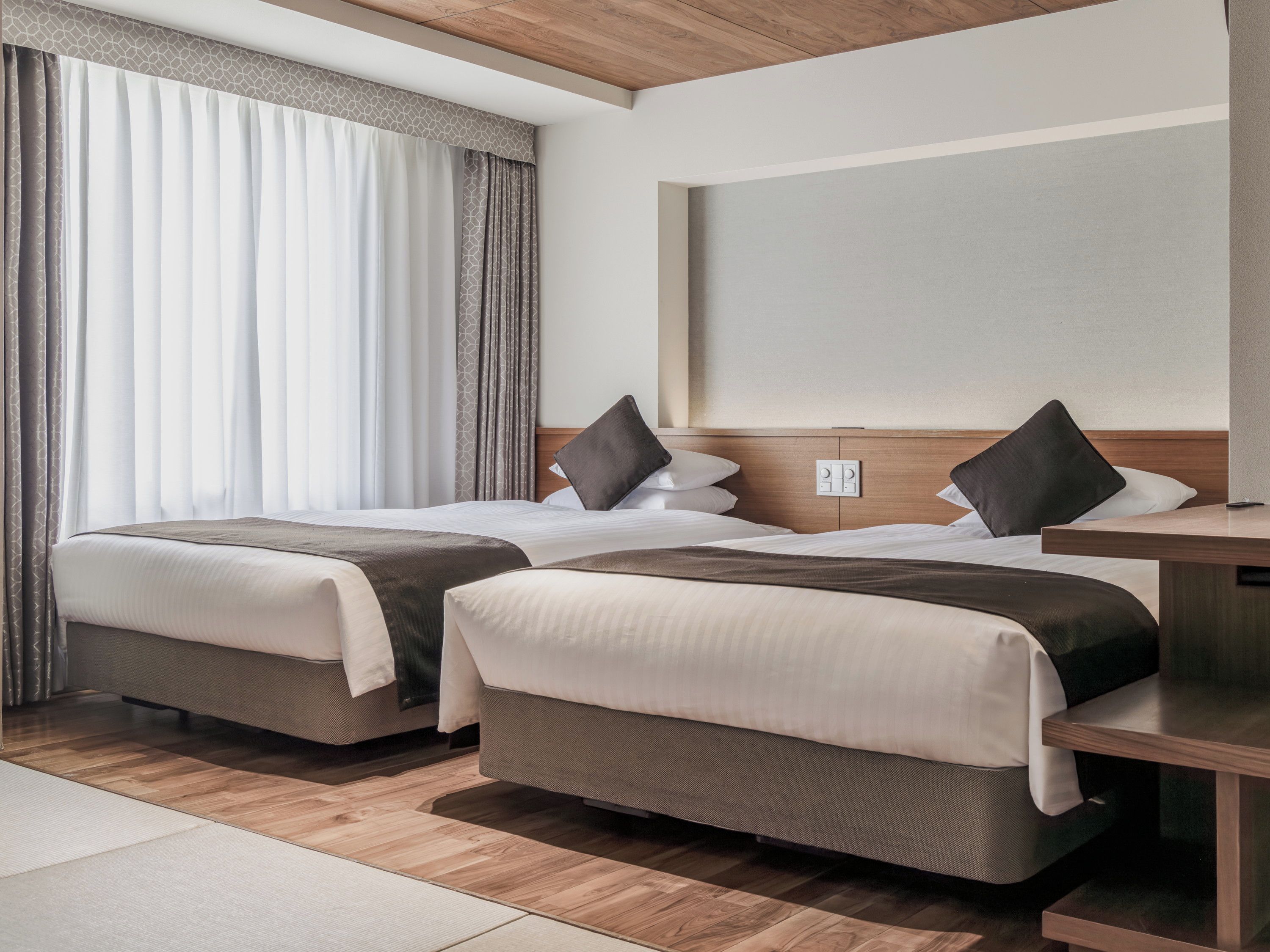 Habitación de hotel con dos camas, cabecero de madera, mesita de noche y ventana con cortinas.