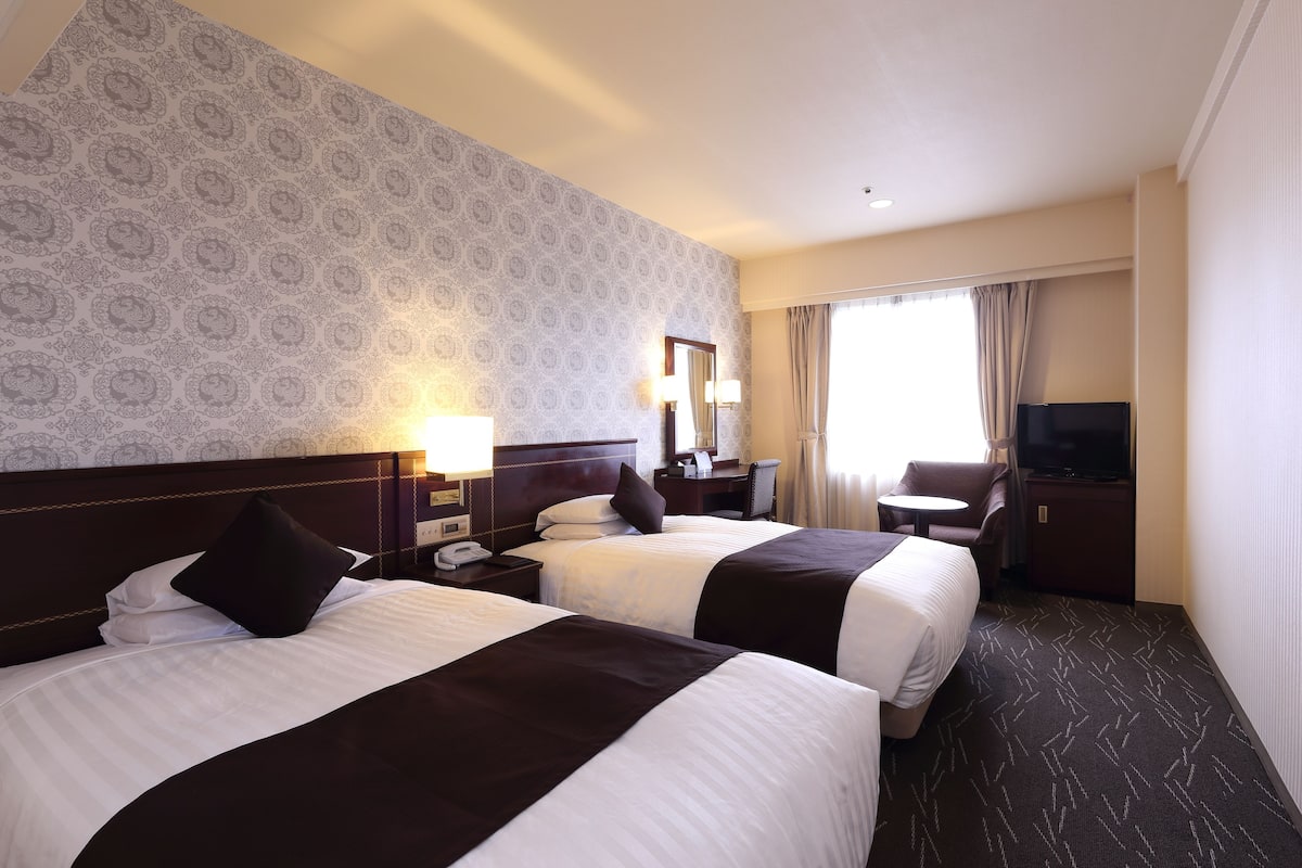 A hotel room with two beds, a desk, a chair, a TV, and a window with curtains.