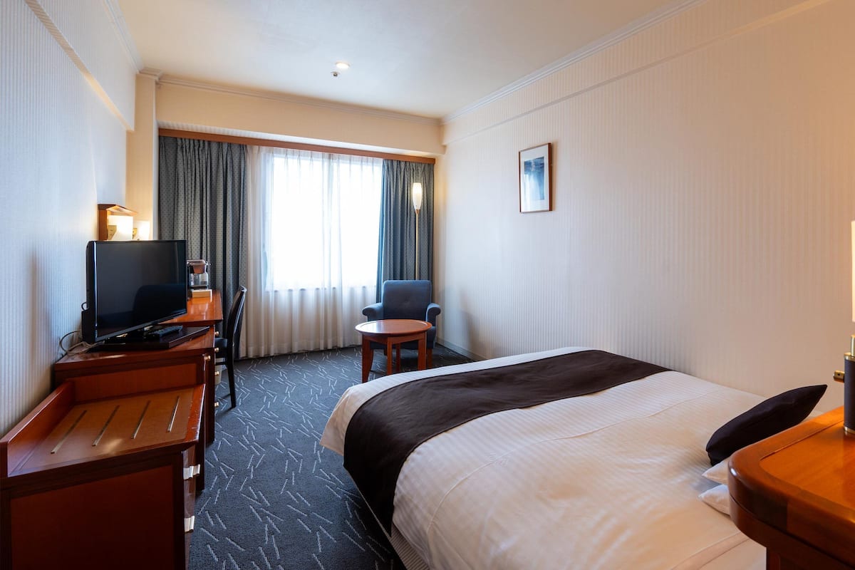 A hotel room with a bed, a desk with a television, a chair, a small table, and a lamp.