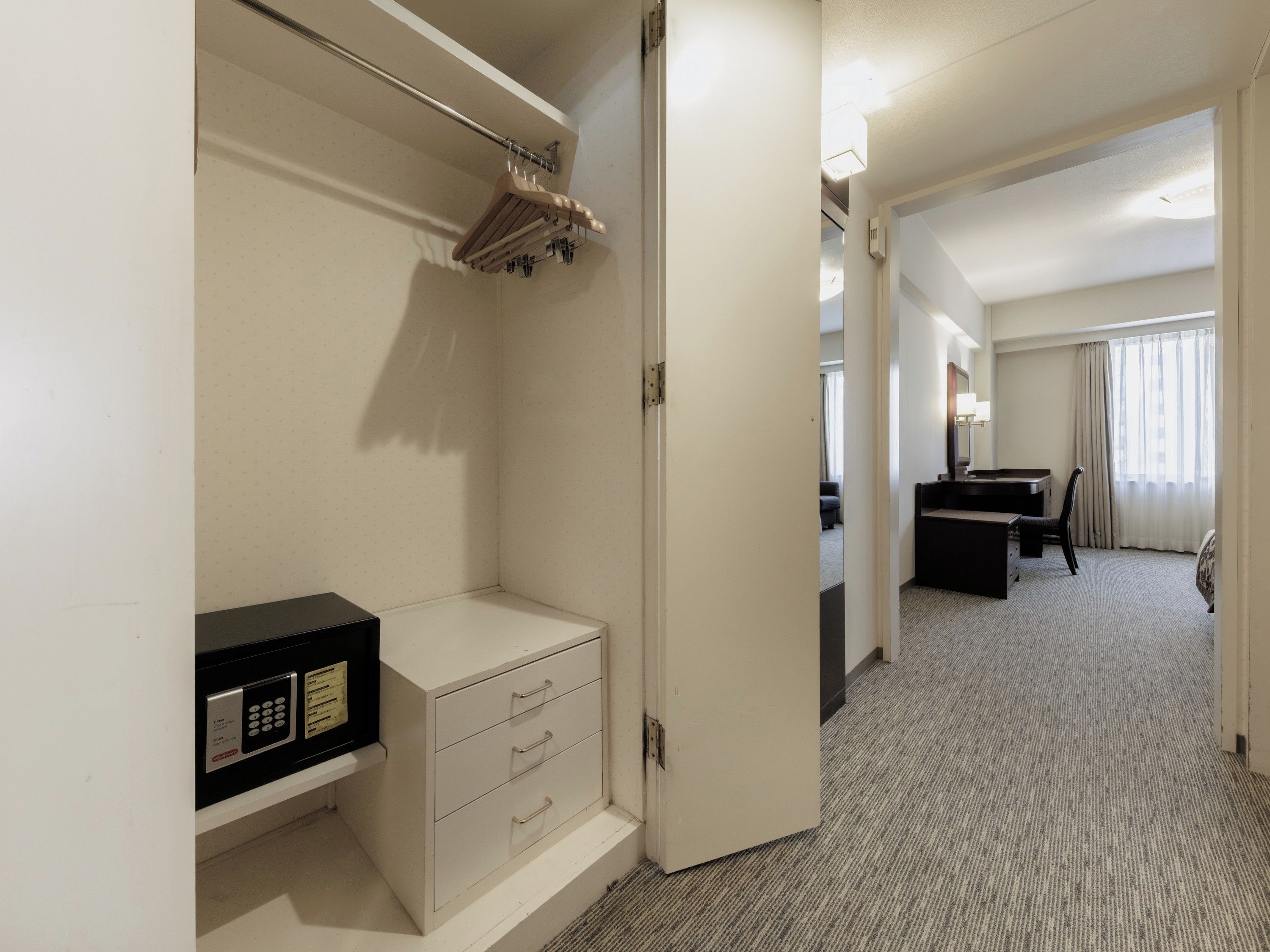 A hotel room with a minibar, a desk, a chair, and a view of the hallway.