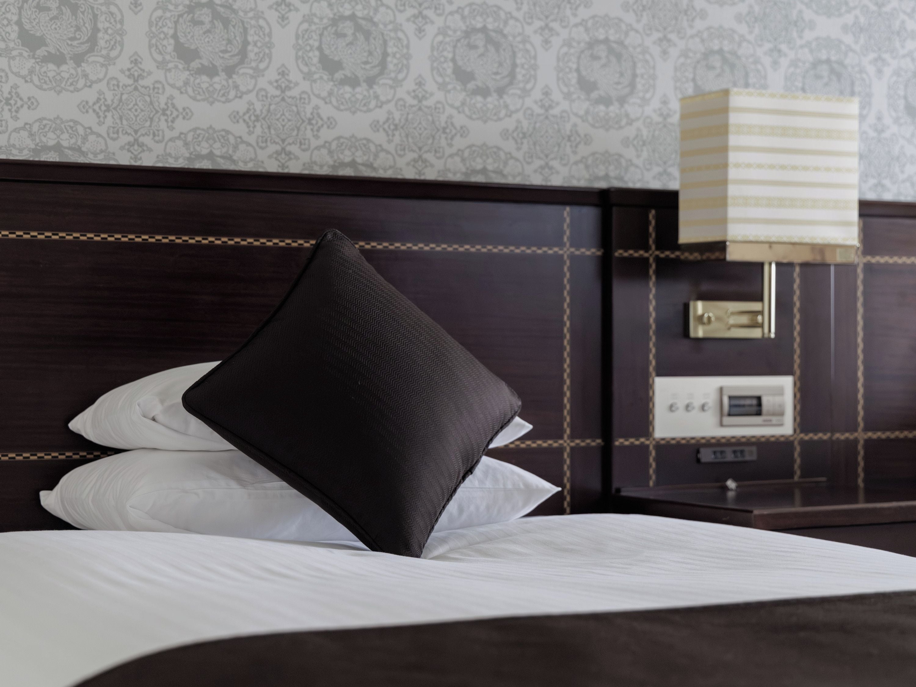 A neatly made bed with white and black pillows, a dark headboard, and a bedside table with a lamp and a thermostat.