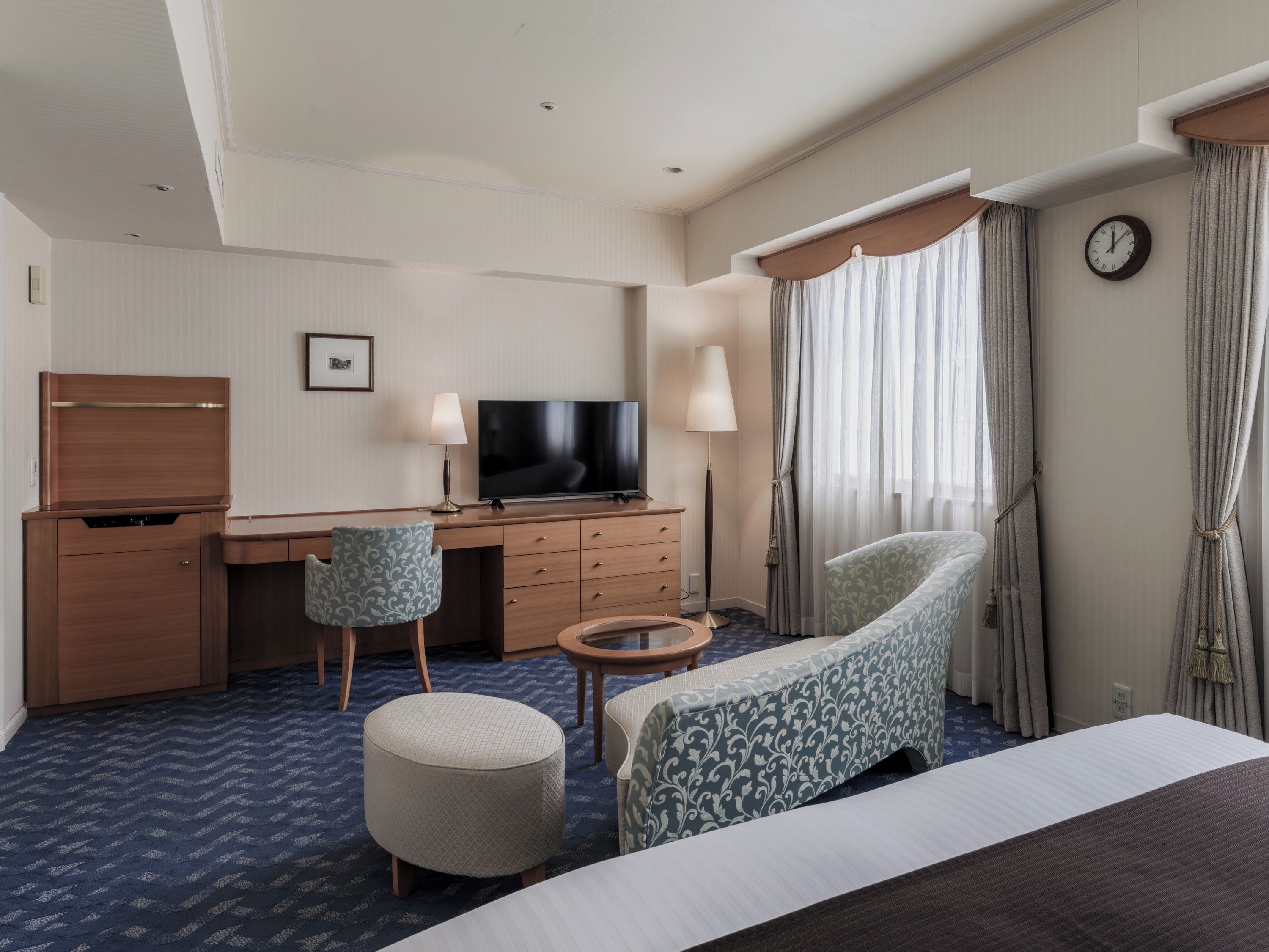 A hotel room with a bed, a desk with a television, a chair, a small table, and a clock on the wall.