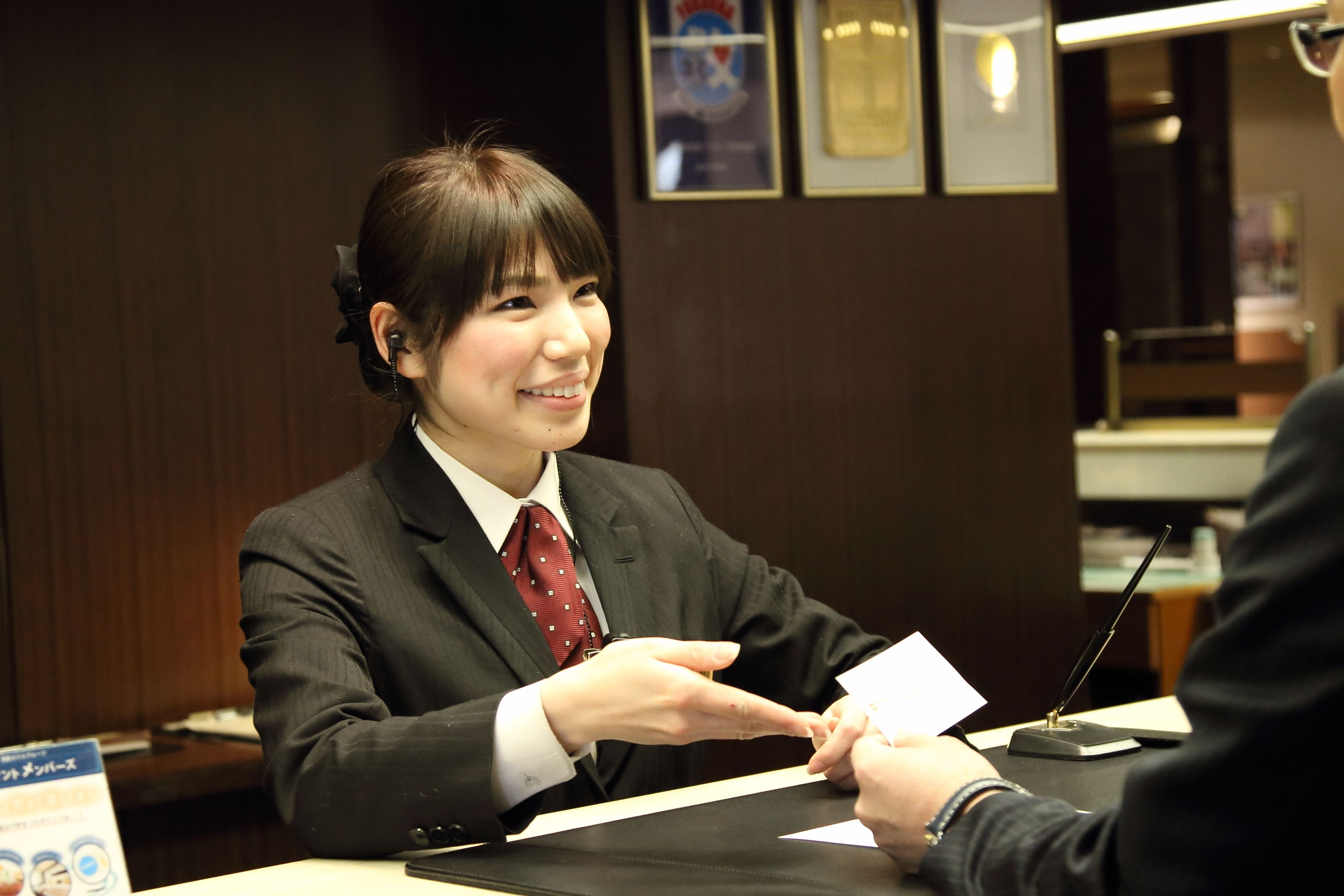 A person in a dark suit and red tie sitting at a table with papers, in a room with framed certificates on the wall.
