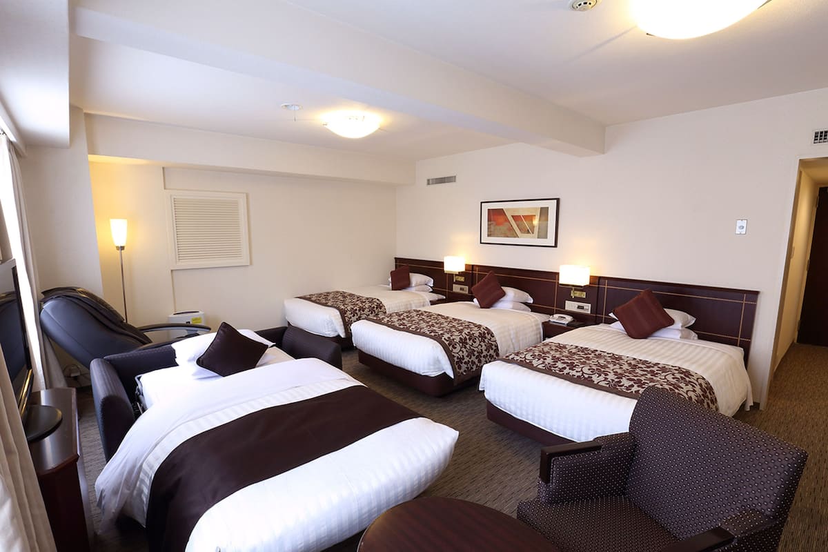 A hotel room with three beds, a desk, a chair, and a lamp.