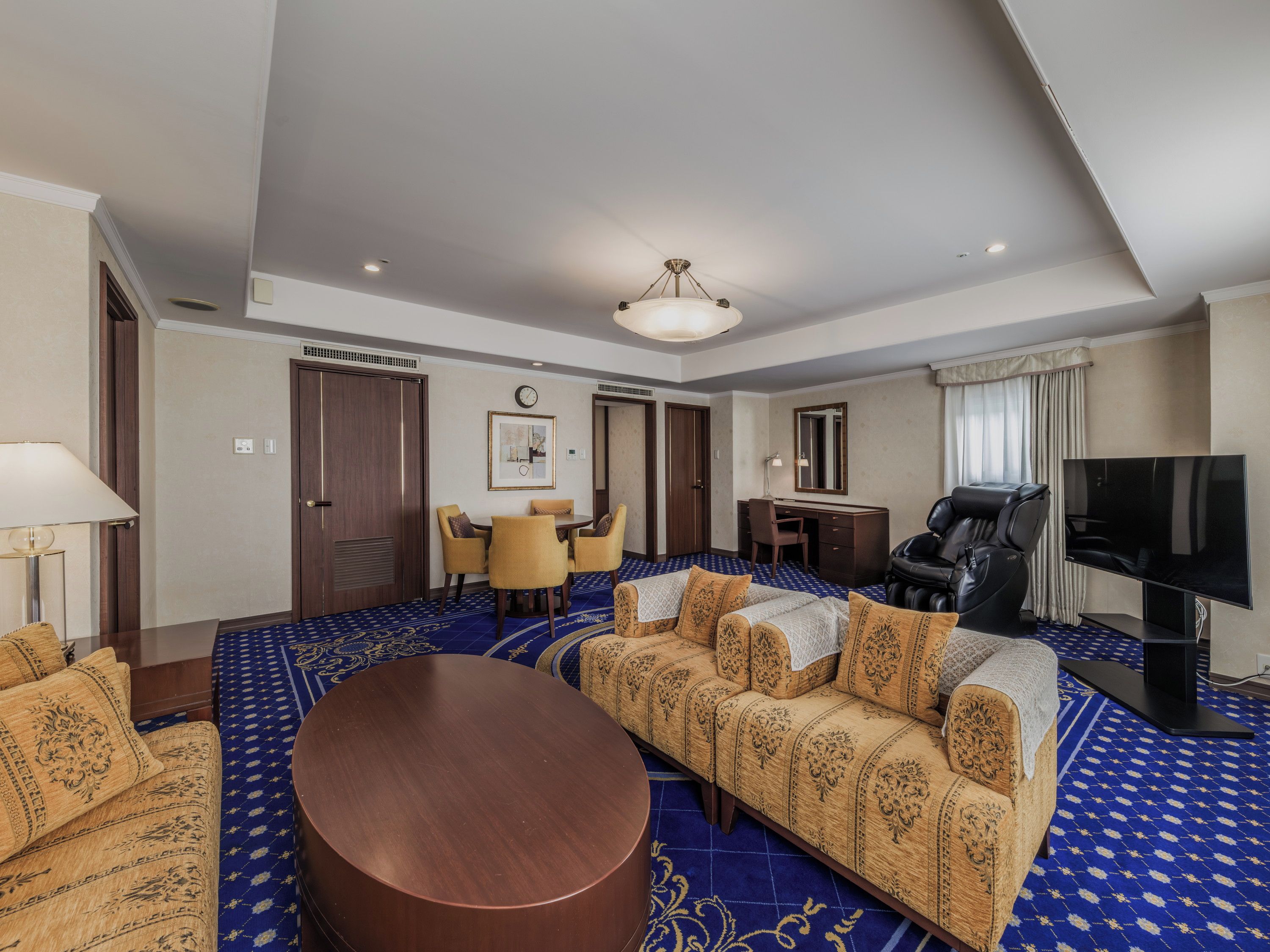 A hotel room with a sofa, a round coffee table, a TV, a desk with a chair, and a dining area with chairs.
