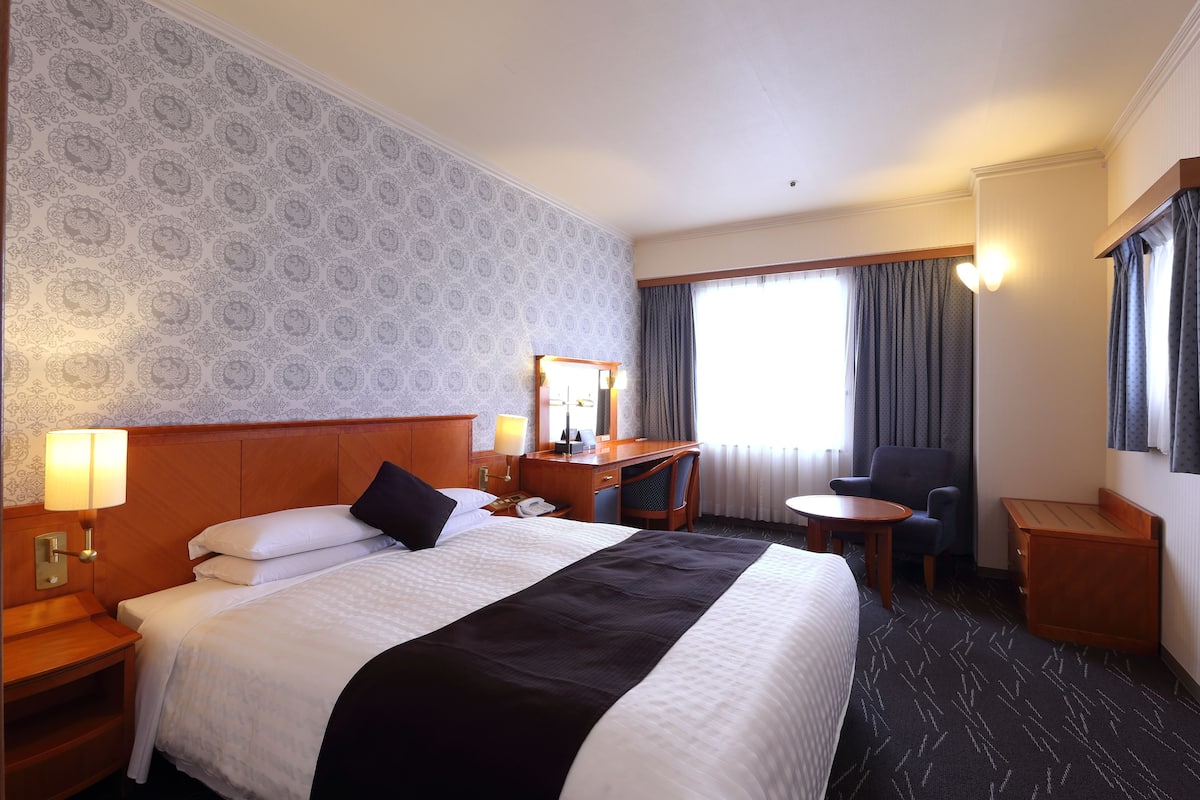 A hotel room with a large bed, a desk, a chair, a small table, and a mirror.