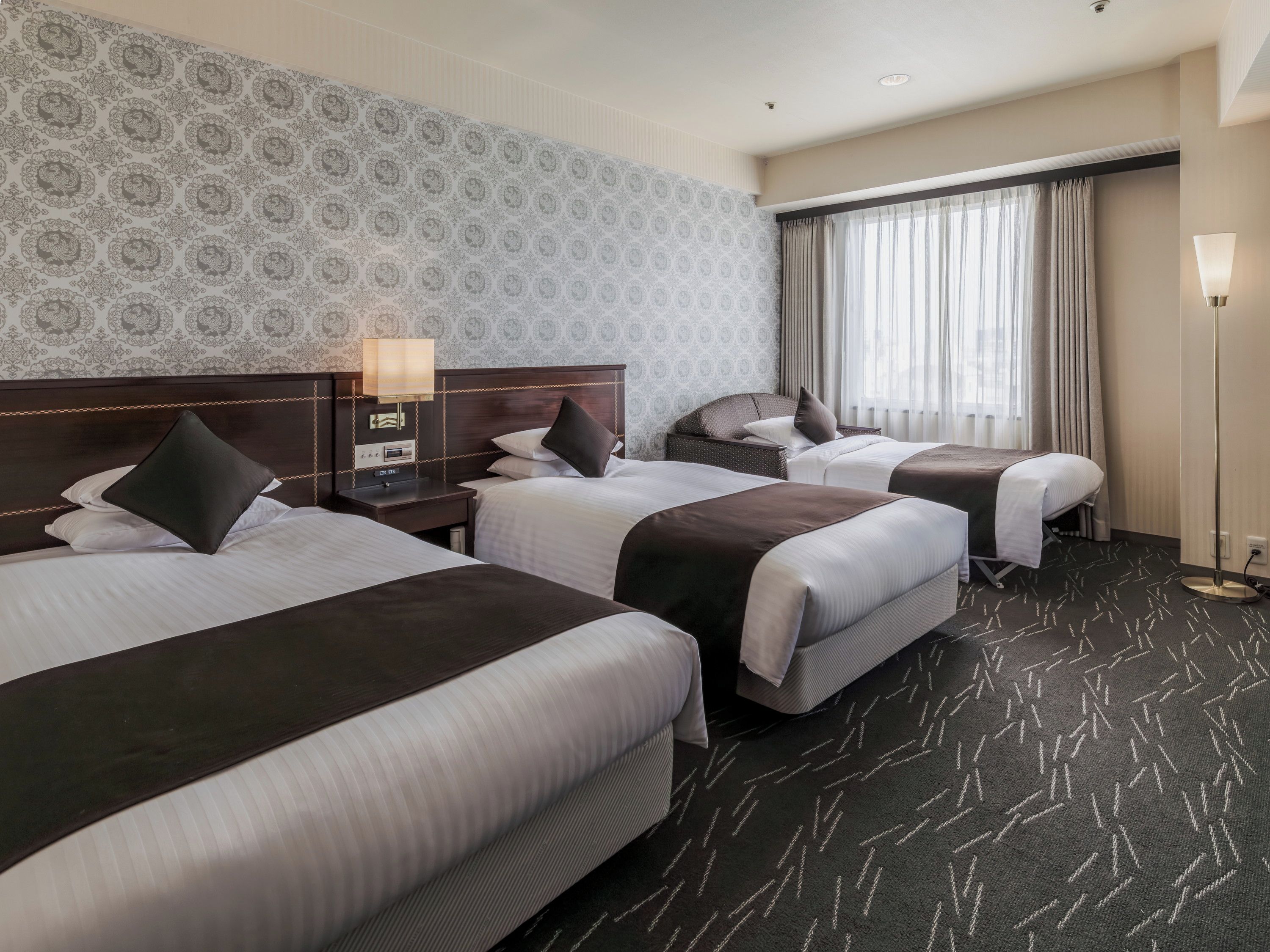 A hotel room with three beds, a patterned wallpaper, a large window with curtains, and a floor lamp.
