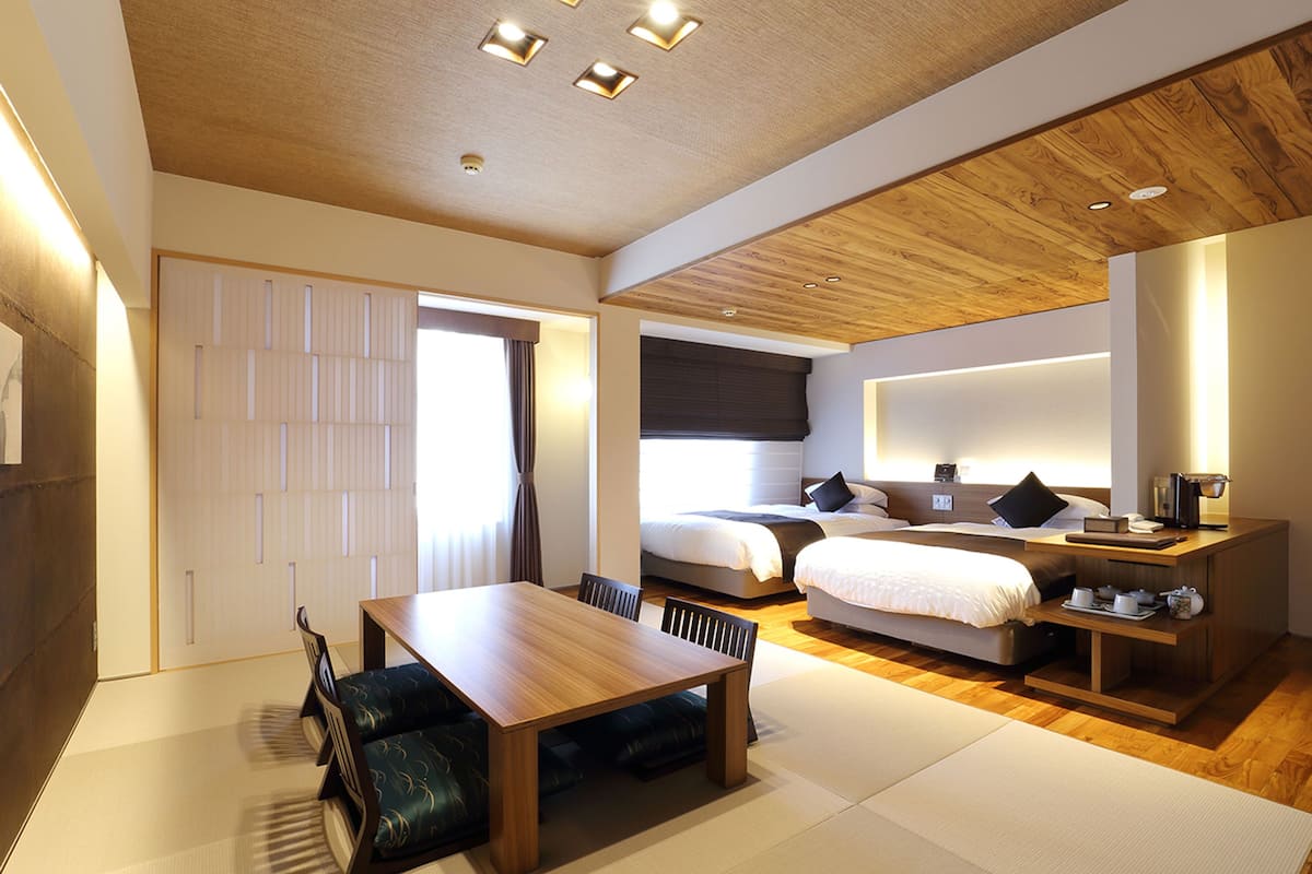 A modern hotel room with a wooden ceiling, a dining table with chairs, a sofa, a bed with a nightstand, and a large window.