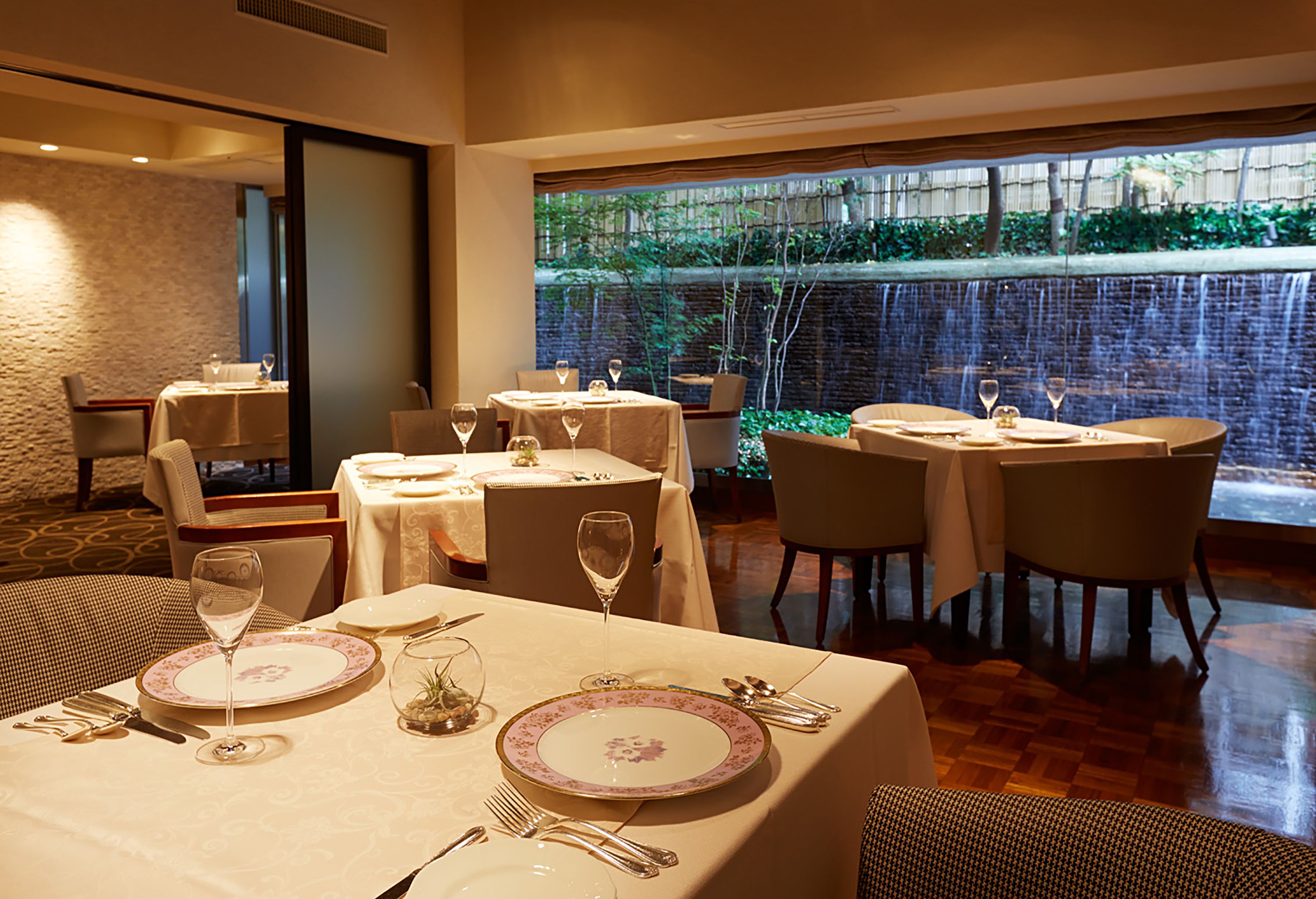 Un restaurante con mesas dispuestas para comer, vista a un jardín a través de un ventanal grande y una pared de ladrillo al fondo.