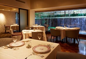 Un restaurante con mesas dispuestas para comer, vista a un jardín a través de un ventanal grande y una pared de ladrillo al fondo.