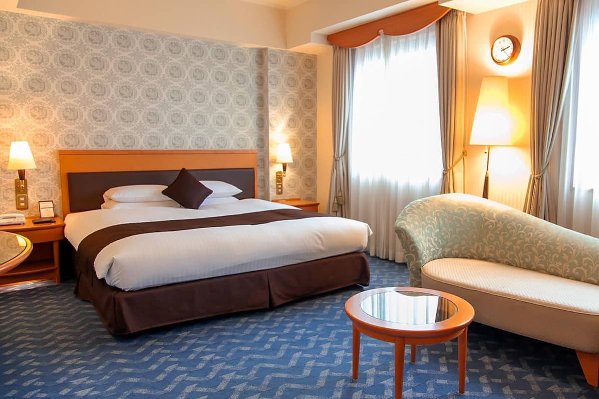 A hotel room with a large bed, two bedside tables with lamps, a sofa, a small round table, and a window with curtains.