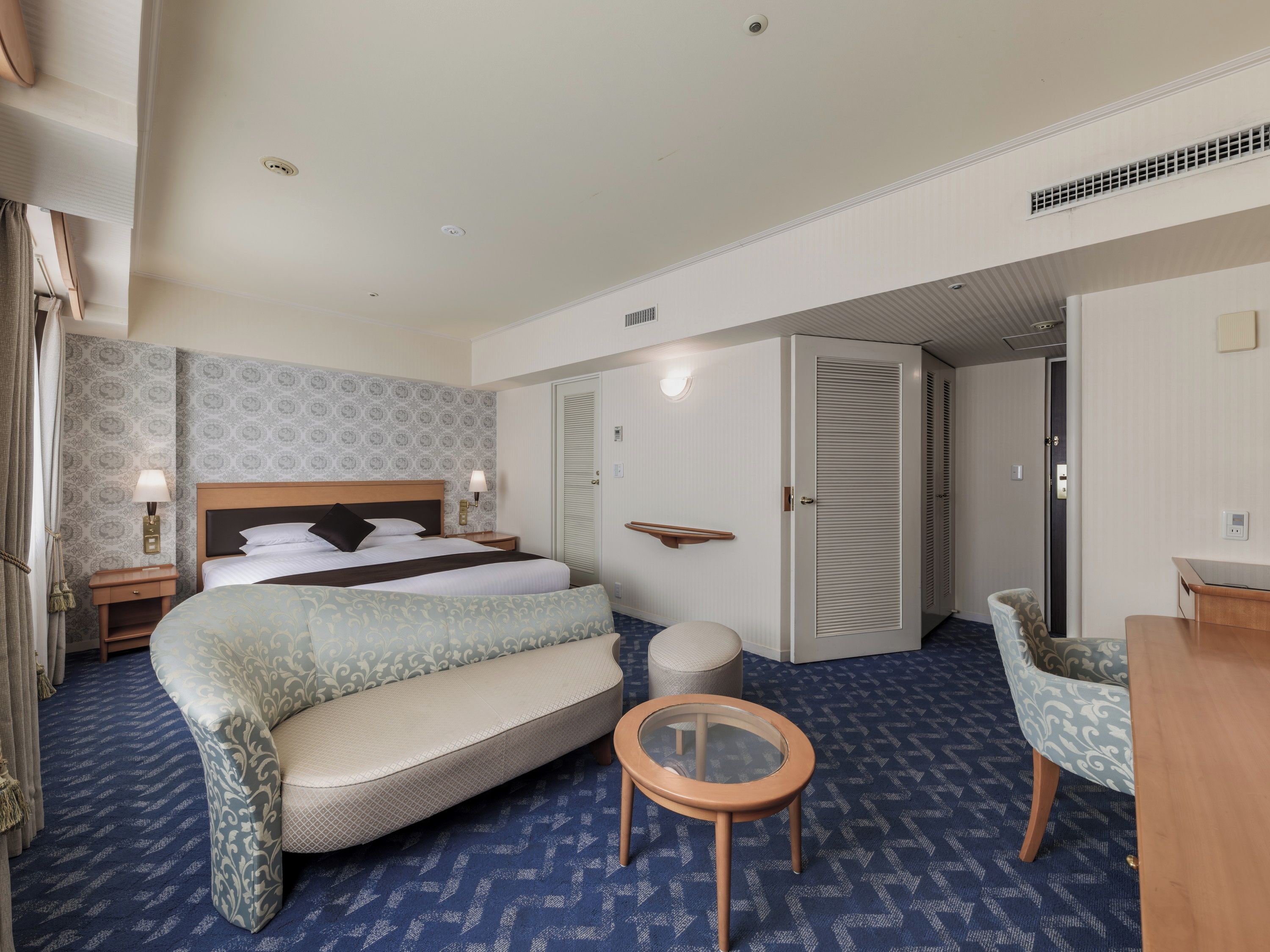 A hotel room with a bed, bedside table, lamp, wardrobe, seating area, and a small round table.
