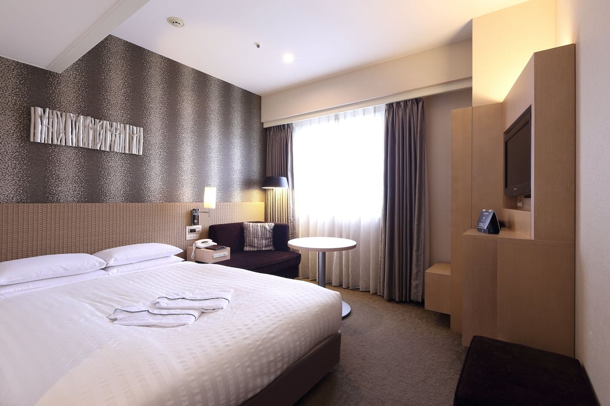 A hotel room with a large bed, a bedside table, a lamp, a TV, and a window with curtains.