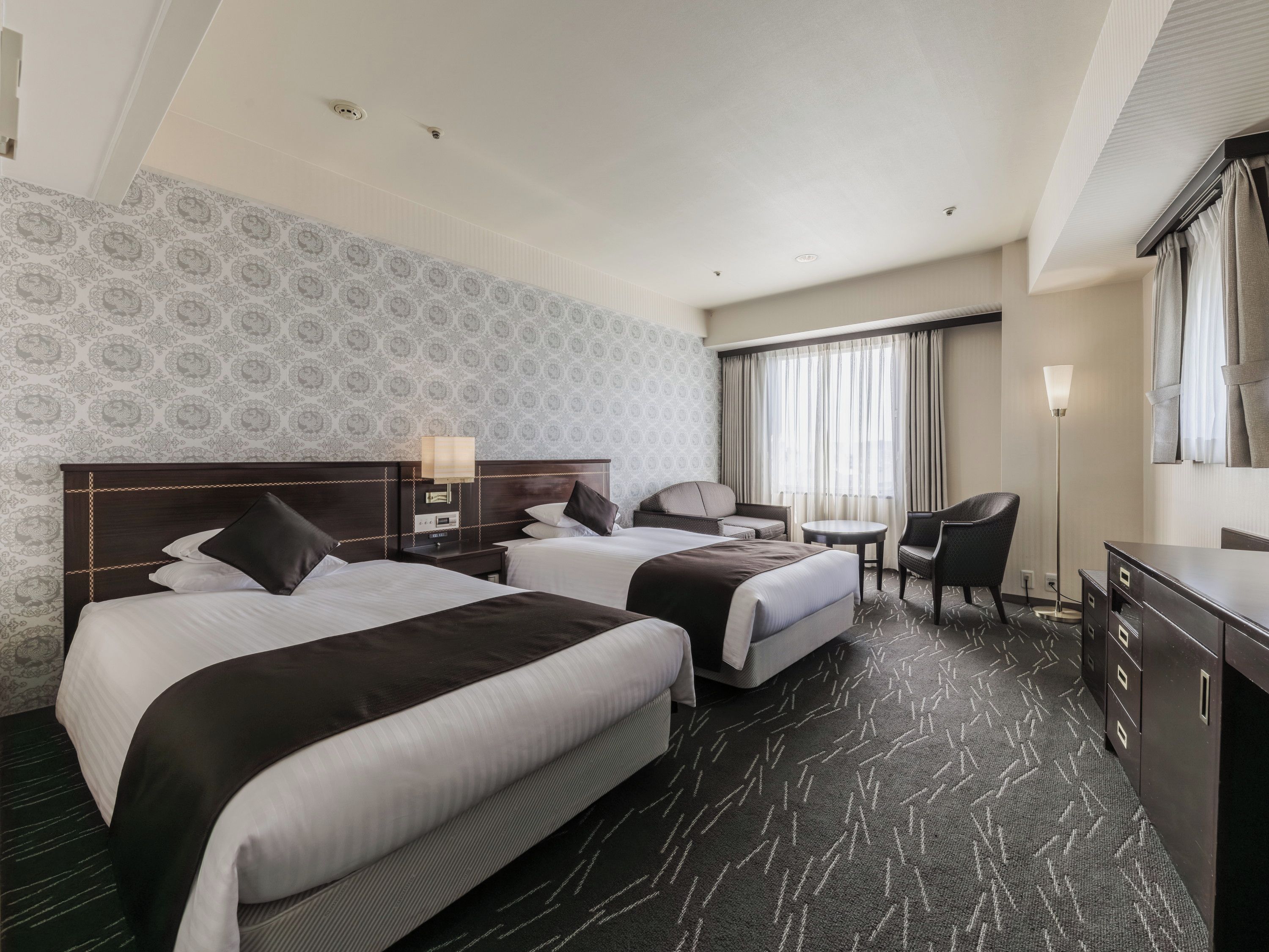 A hotel room with two beds, a desk, a chair, a lamp, and a large window with curtains.