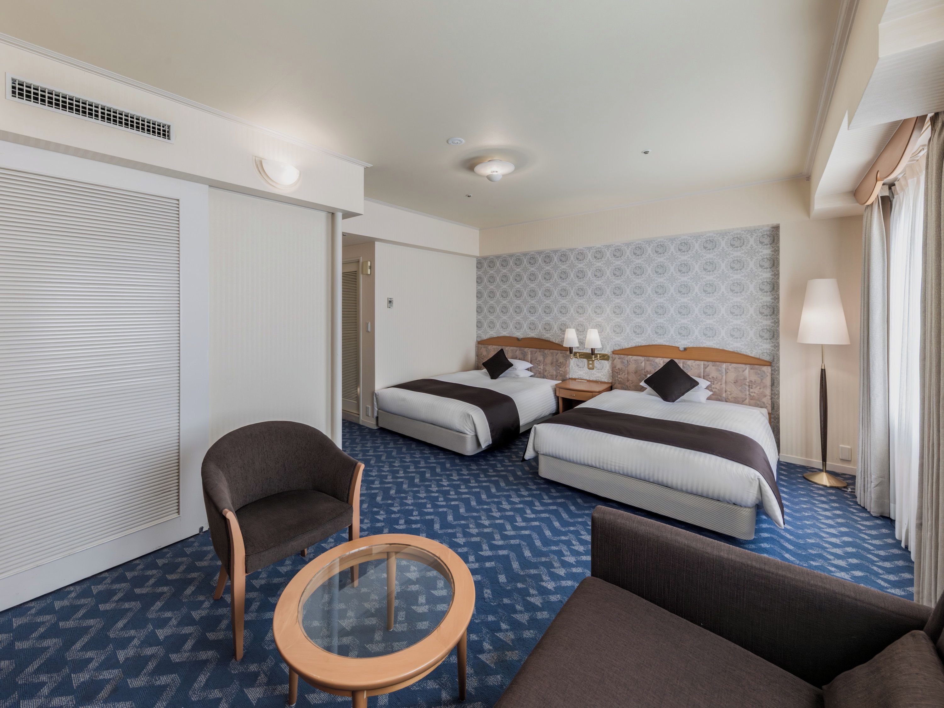 A hotel room with two beds, a seating area with a chair and a small table, a lamp, and a patterned carpet.