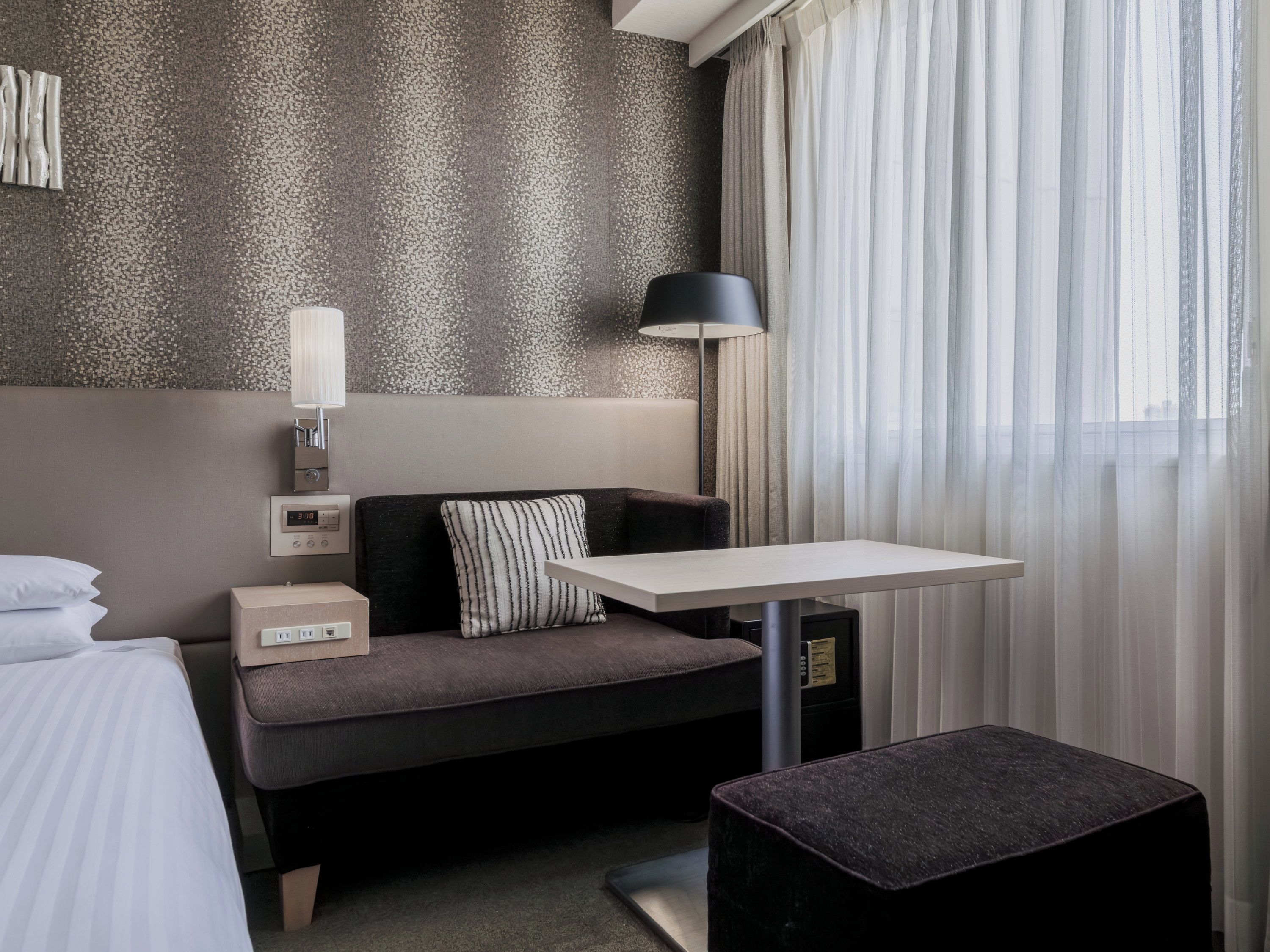 Hotellihuone, jossa on sänky, työpöytä, tuoli, yöpöytä, lamppu ja ikkuna, jossa on verhot.