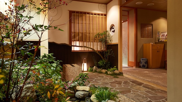 Un interior japonés tradicional con un sendero de piedra que lleva a una puerta de madera, rodeado de abundante vegetación y un farol.