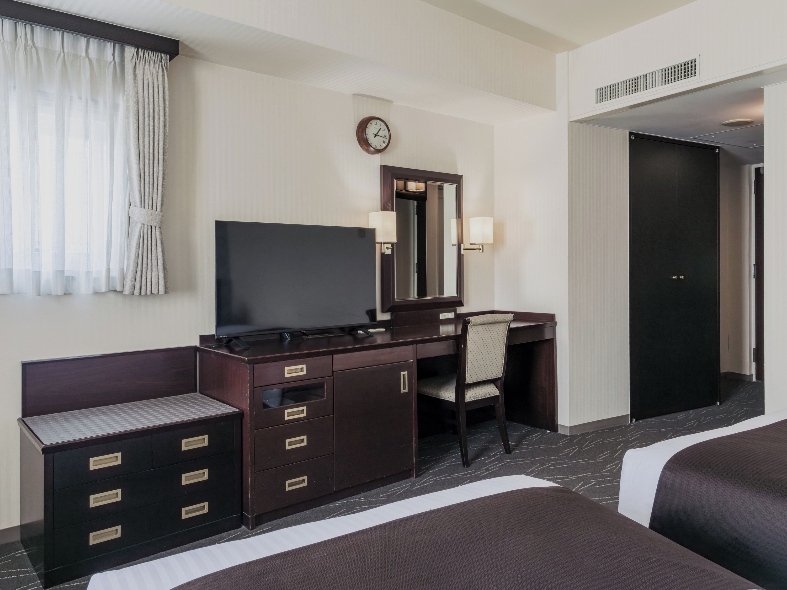 A hotel room with a bed, a desk with a flat-screen TV, a chair, a dresser, a clock, and a door.