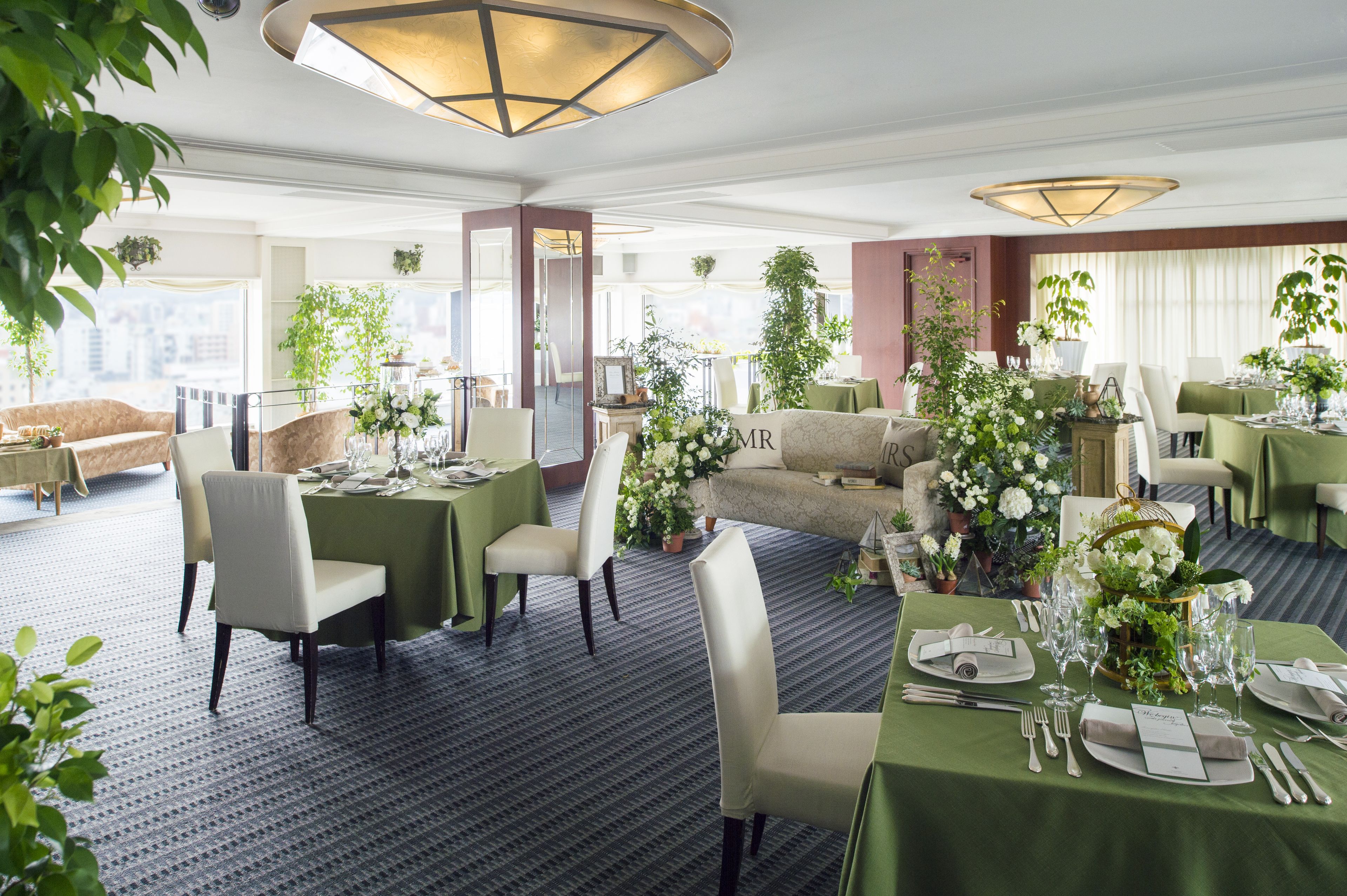 Comedor de hotel con mesas dispuestas para una comida, rodeado de asientos tapizados en tonos verdes y plantas en macetas.