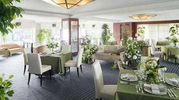 Comedor de hotel con mesas dispuestas para una comida, rodeado de asientos tapizados en tonos verdes y plantas en macetas.