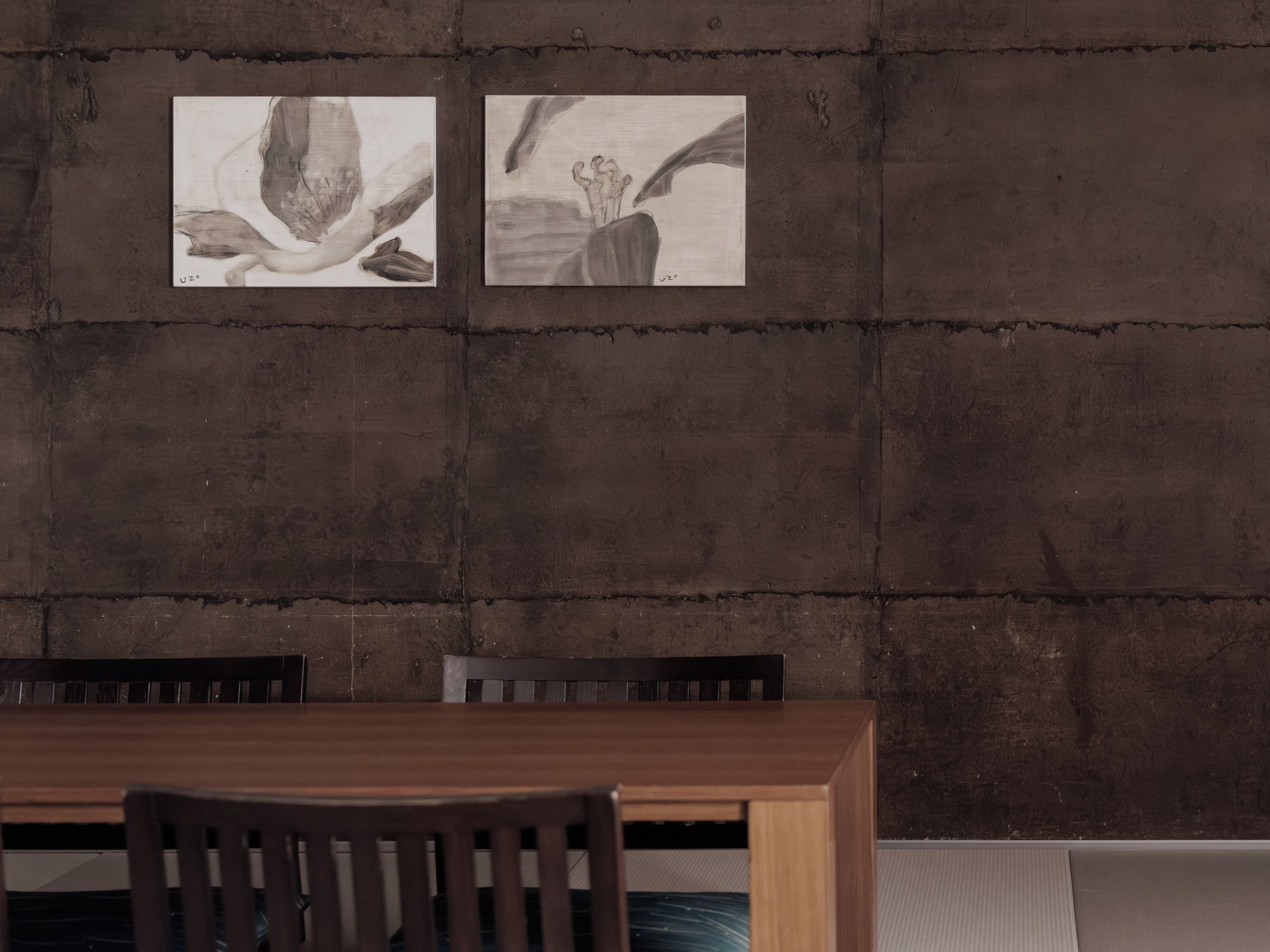 A dining area with a wooden table and chairs, a concrete wall with two framed artworks, and a textured floor.