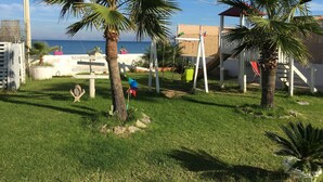 A grassy outdoor area with palm trees, a swing set, and a view of the sea. - Case Playa (Balestrate)