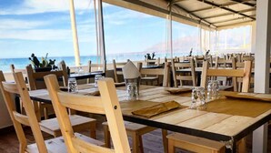 A restaurant with wooden tables and chairs, large windows offering a view of the sea, and a covered outdoor area. - Case Playa (Balestrate)