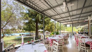 Un restaurante al aire libre con mesas cubiertas de manteles a cuadros rojos y blancos, sillas blancas y vistas a una piscina y zonas verdes.