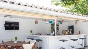 Un patio al aire libre con una mesa redonda de madera, cuatro taburetes altos y una barra blanca con taburetes.
