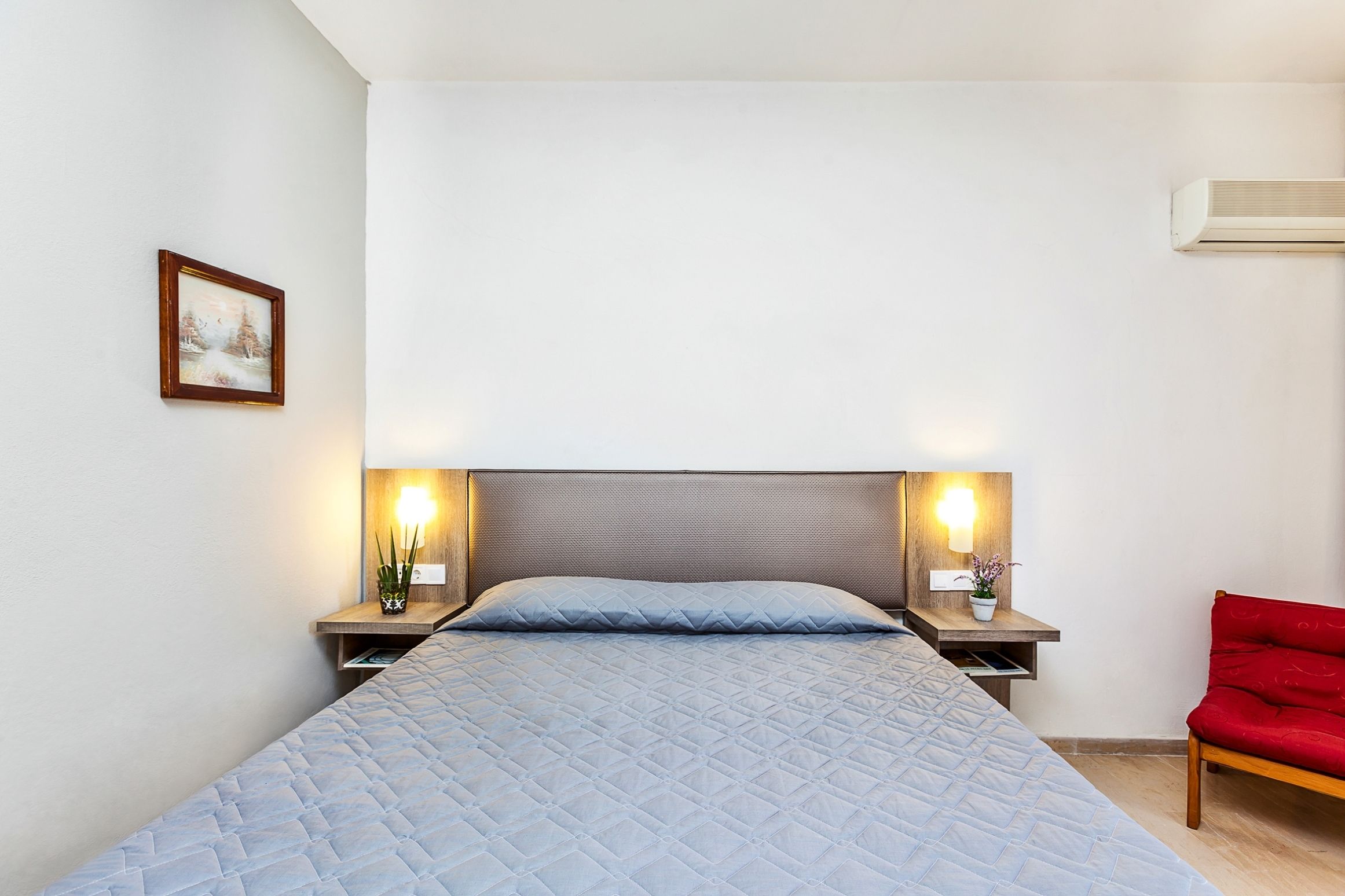 A bed with a quilted blue cover, two bedside tables with lamps, a red chair, and a wall-mounted air conditioner.