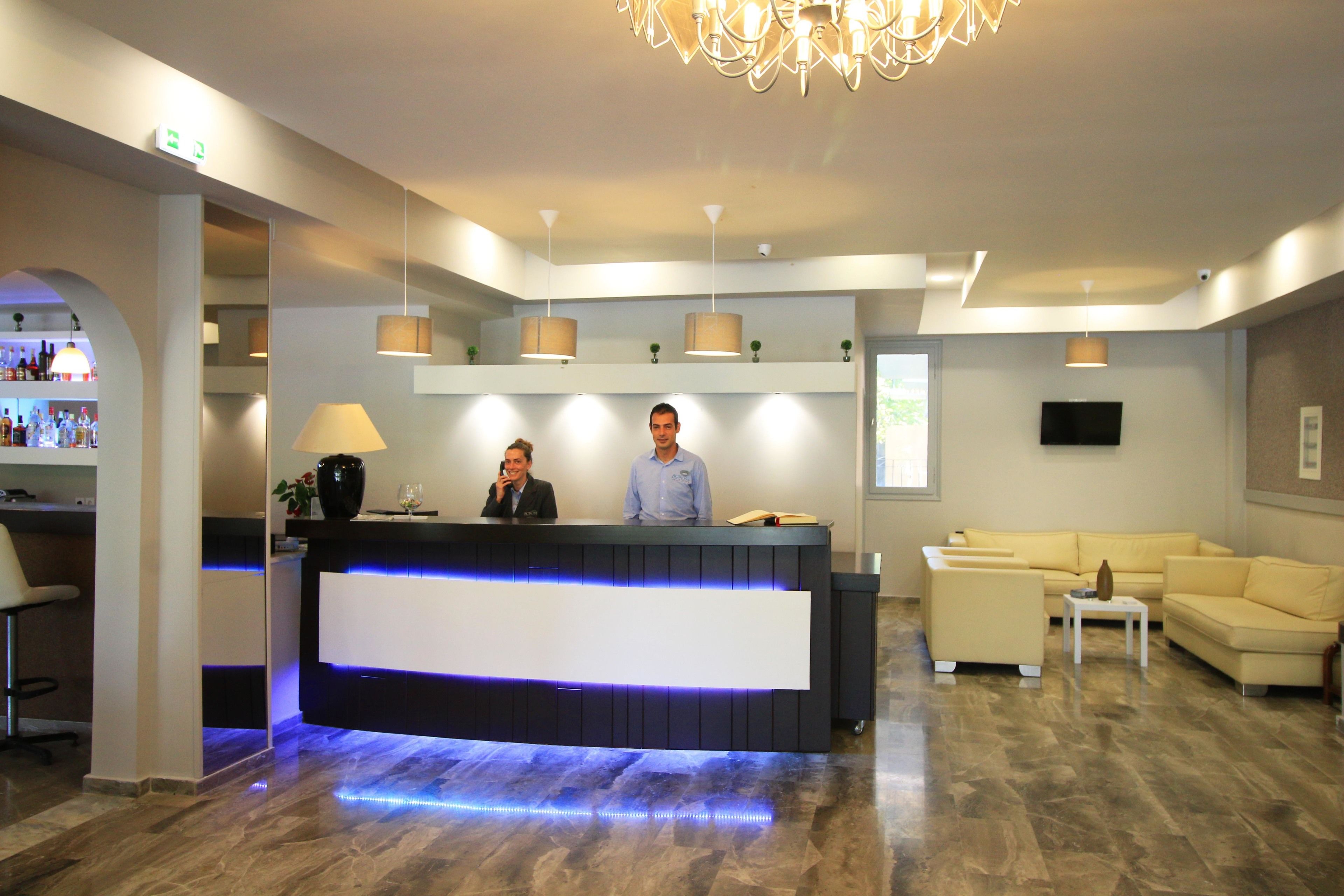 A modern reception area with a reception desk, two staff members, a bar area, and a seating area with a yellow sofa and a small table.