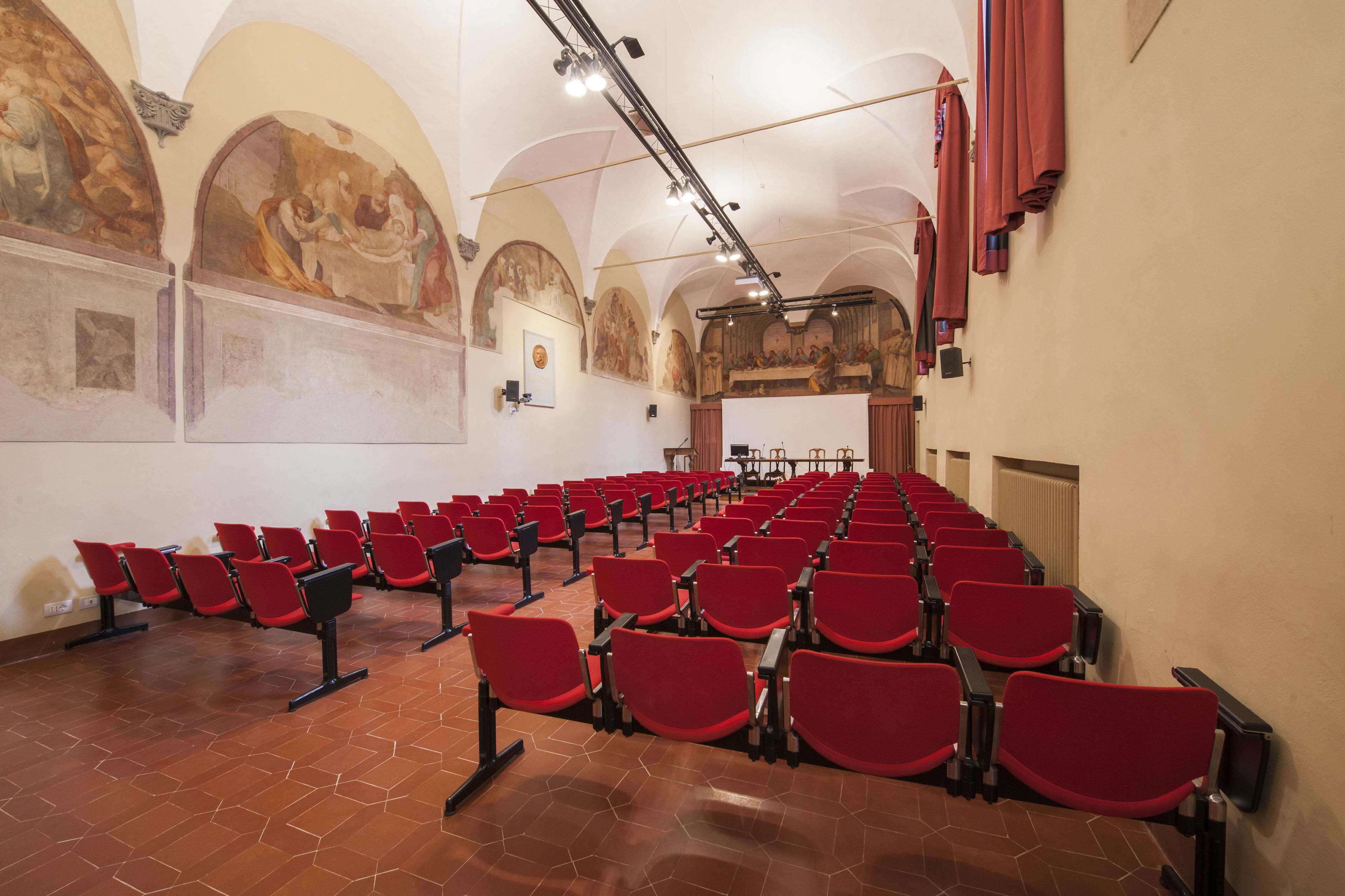 Een ruime zaal met rijen rode stoelen, hoge gewelfde plafonds en klassieke fresco's op de muren.