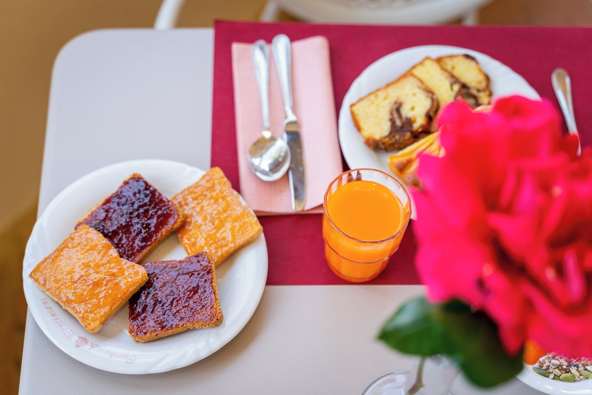 Ein Frühstückstisch mit Toast, Marmelade, einem Glas Orangensaft und einer rosa Blume.