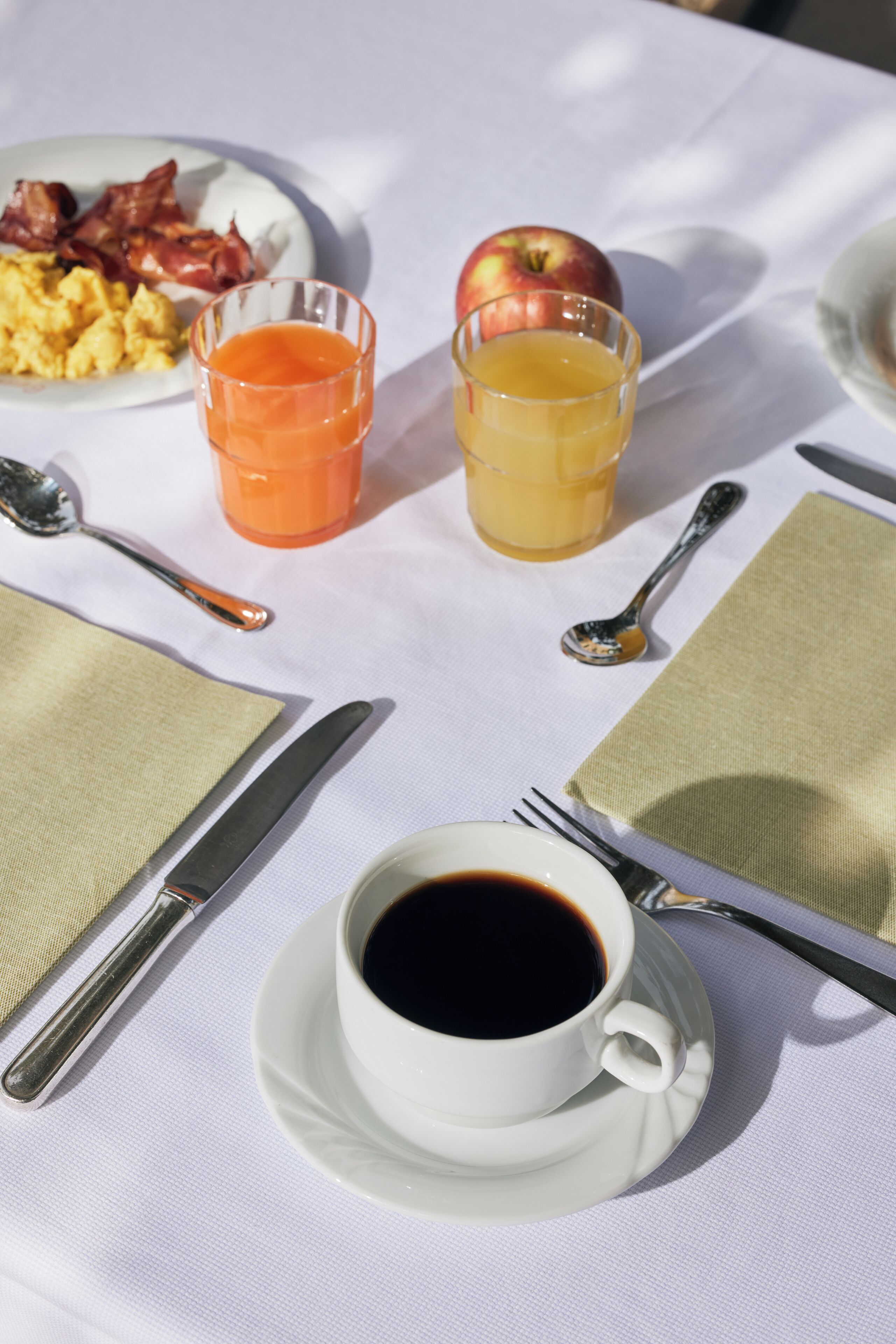A breakfast table with a plate of scrambled eggs, bacon, an apple, orange juice, and coffee.