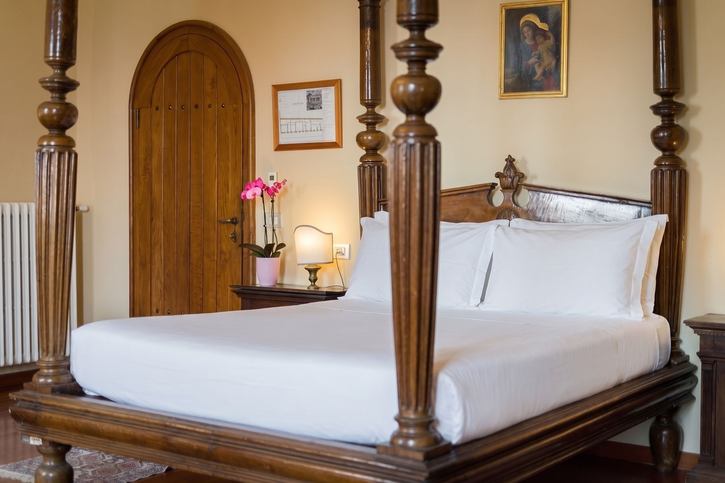 Un letto a baldacchino con struttura in legno e biancheria bianca, una porta in legno, un certificato incorniciato appeso al muro e una pianta in vaso con fiori rosa.