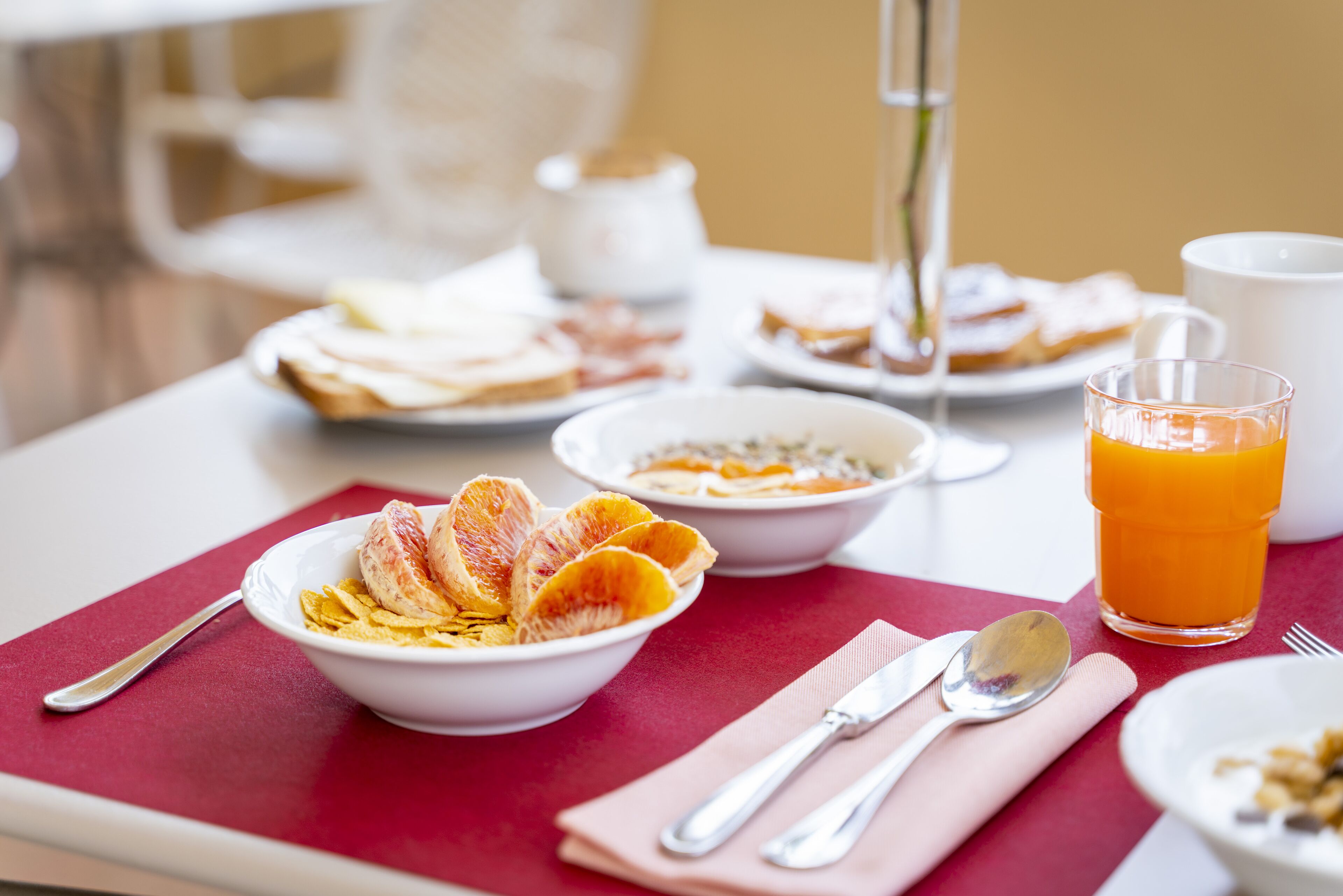 A breakfast setting with a table set for two, including plates of cereal, toast, and orange slices, a glass of orange juice, and a vase with a plant.