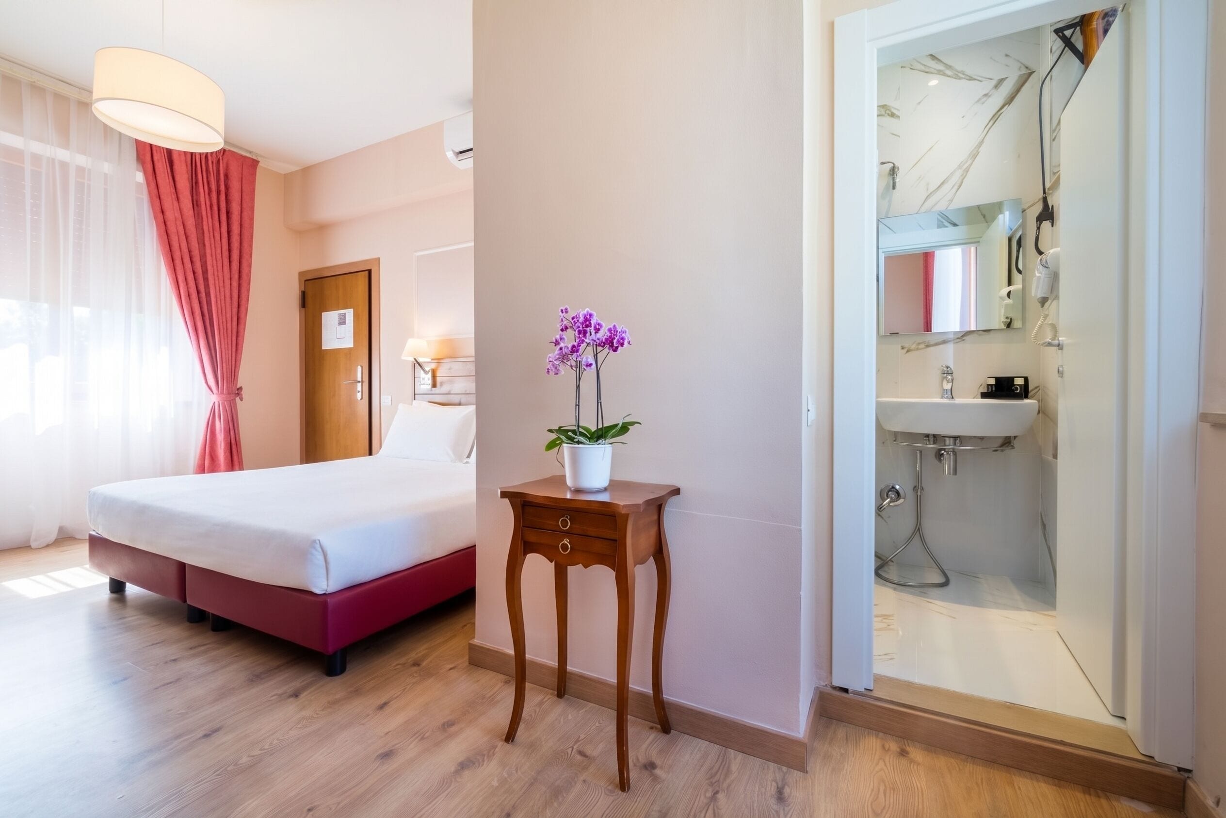 A hotel room with a bed, a bedside table with a plant, a bathroom with a shower, and a mirror.