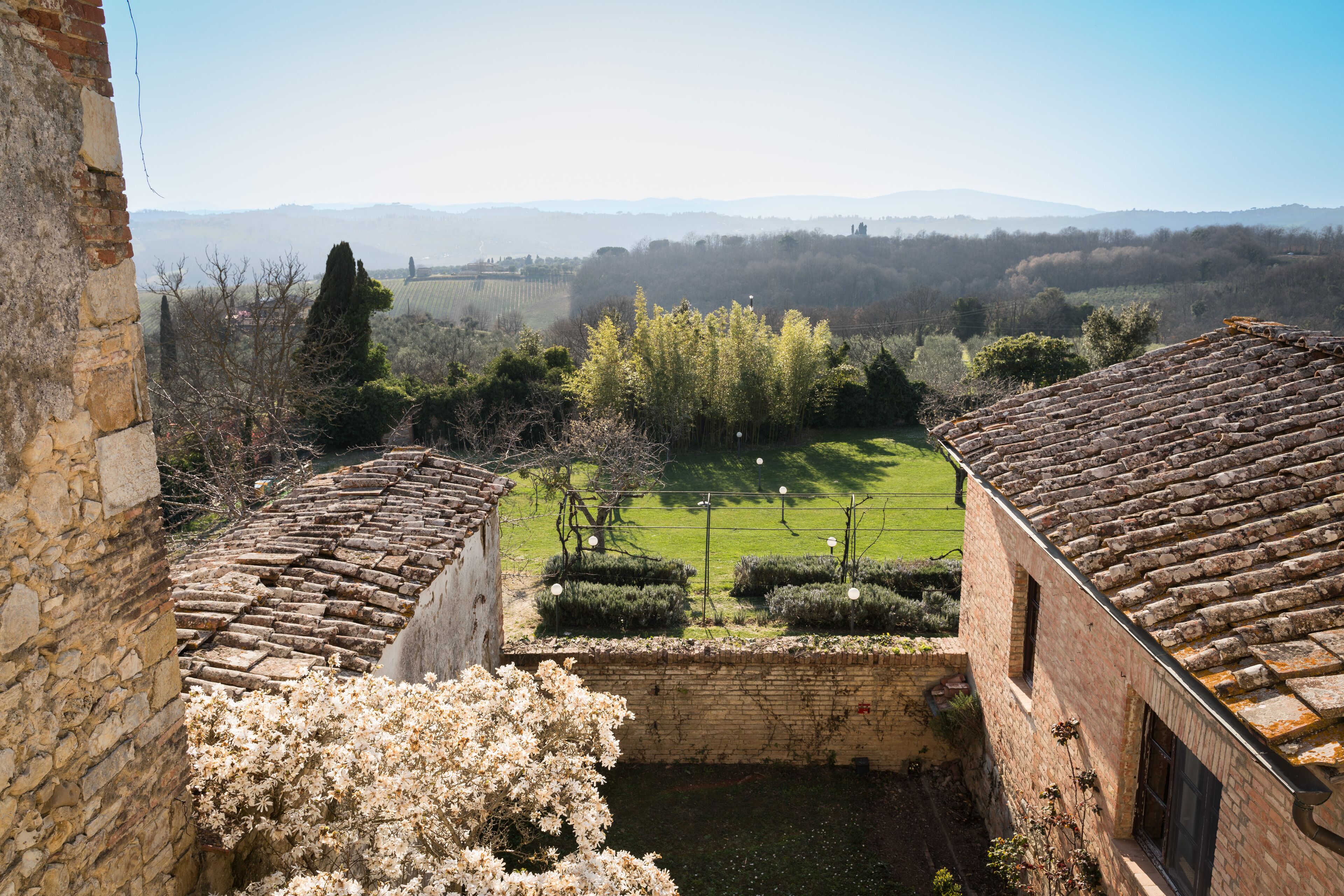 Ein Innenhof mit Blick auf ein grünes Feld und ferne Hügel, umgeben von Steingebäuden mit Terrakotta-Dächern.