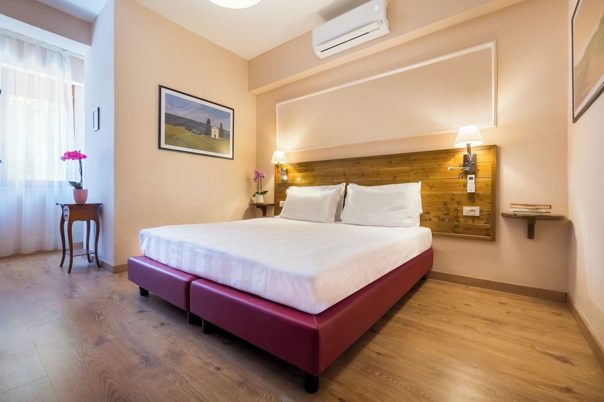 Habitación de hotel con cabecera de madera, cama con ropa blanca, mesita de noche con lámpara y una pequeña planta en maceta.