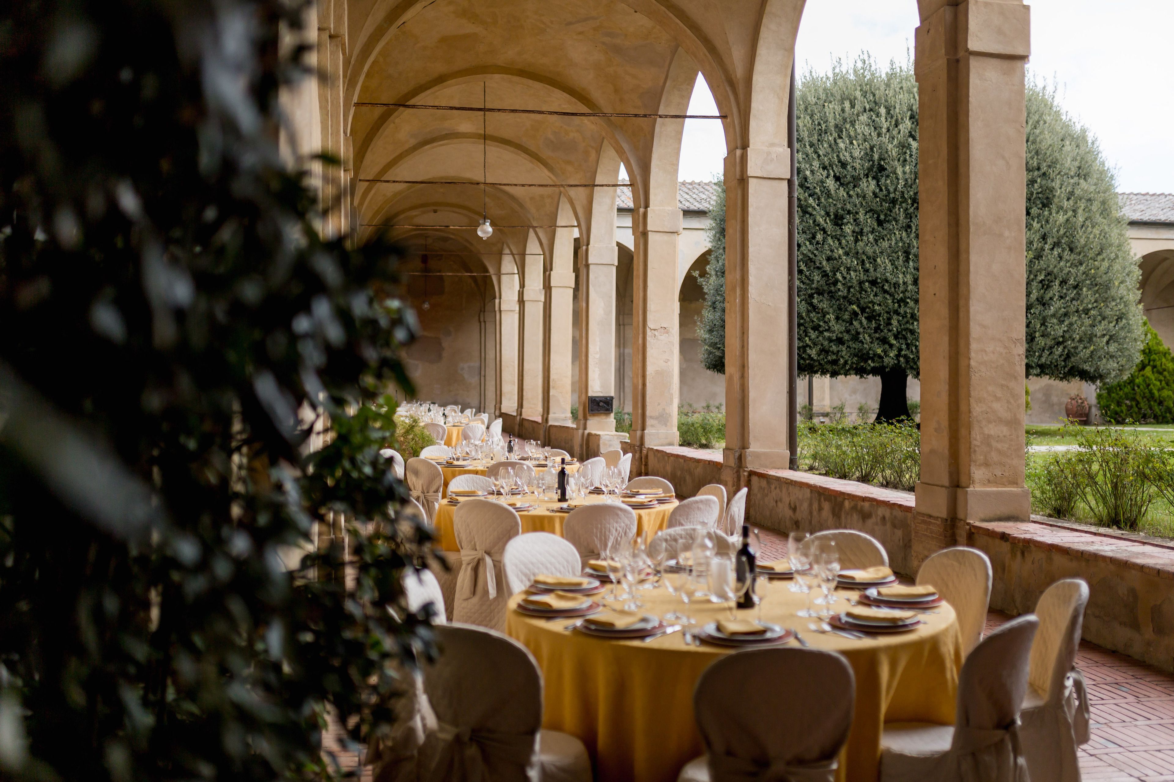 Un espace de restauration en plein air avec des tables rondes dressées pour un repas, entouré d’arcades et de verdure.